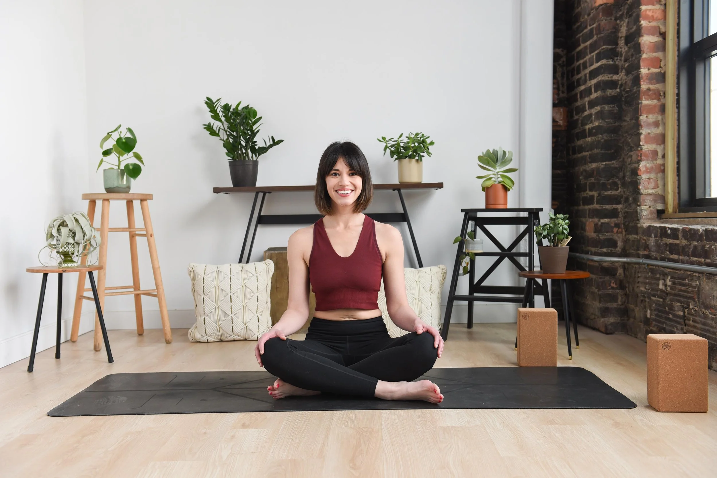 Woman practicing yoga indoors, sitting cross-legged on a yoga mat, smiling, with a background of potted plants and a brick wall.