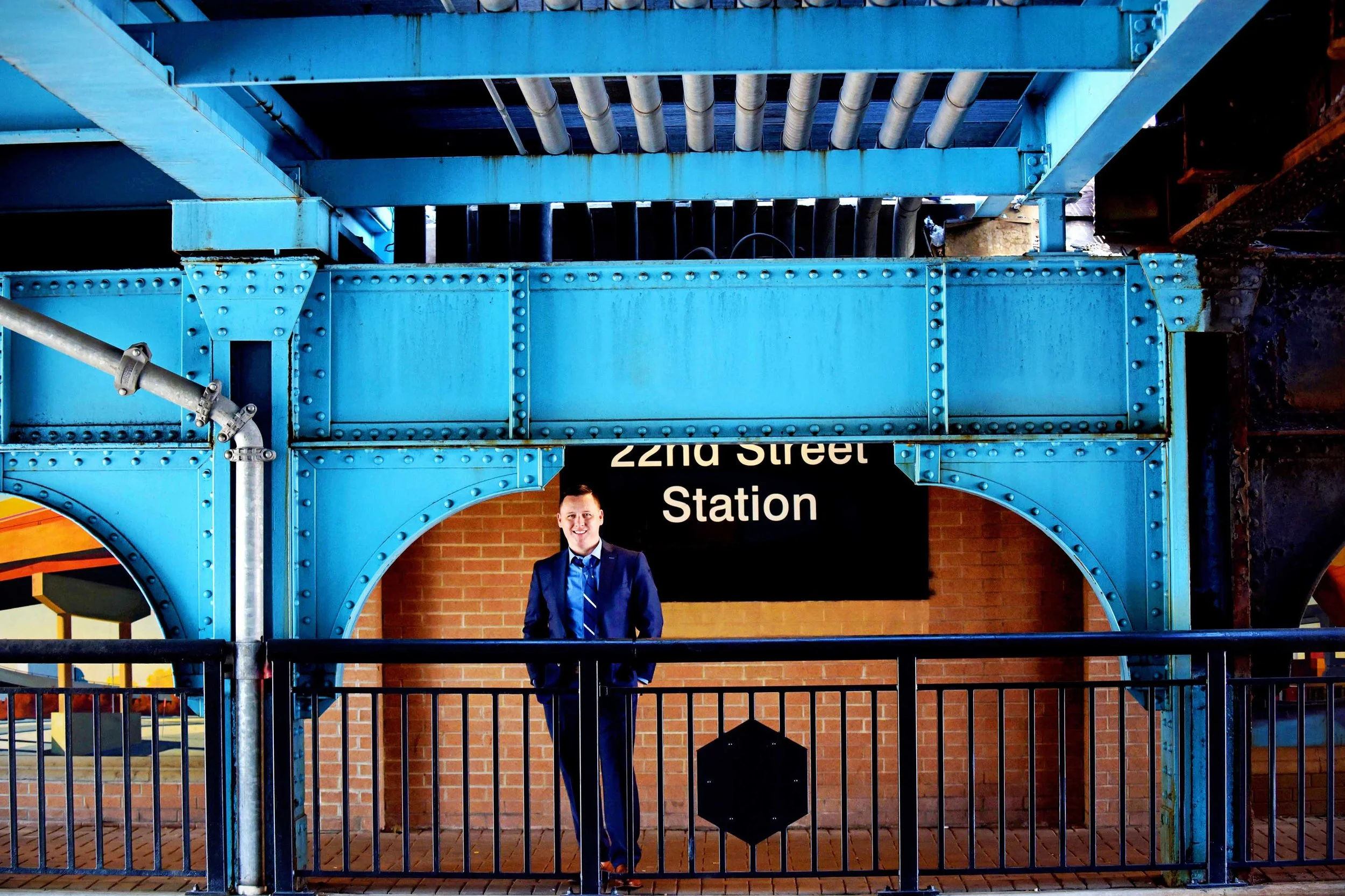 A man in a blue suit and striped tie standing in front of a brick wall and a black sign that reads '22nd Street Station'. The man is inside a blue metal structure with pipes and rivets, and the bottom part of the image shows a black railing.