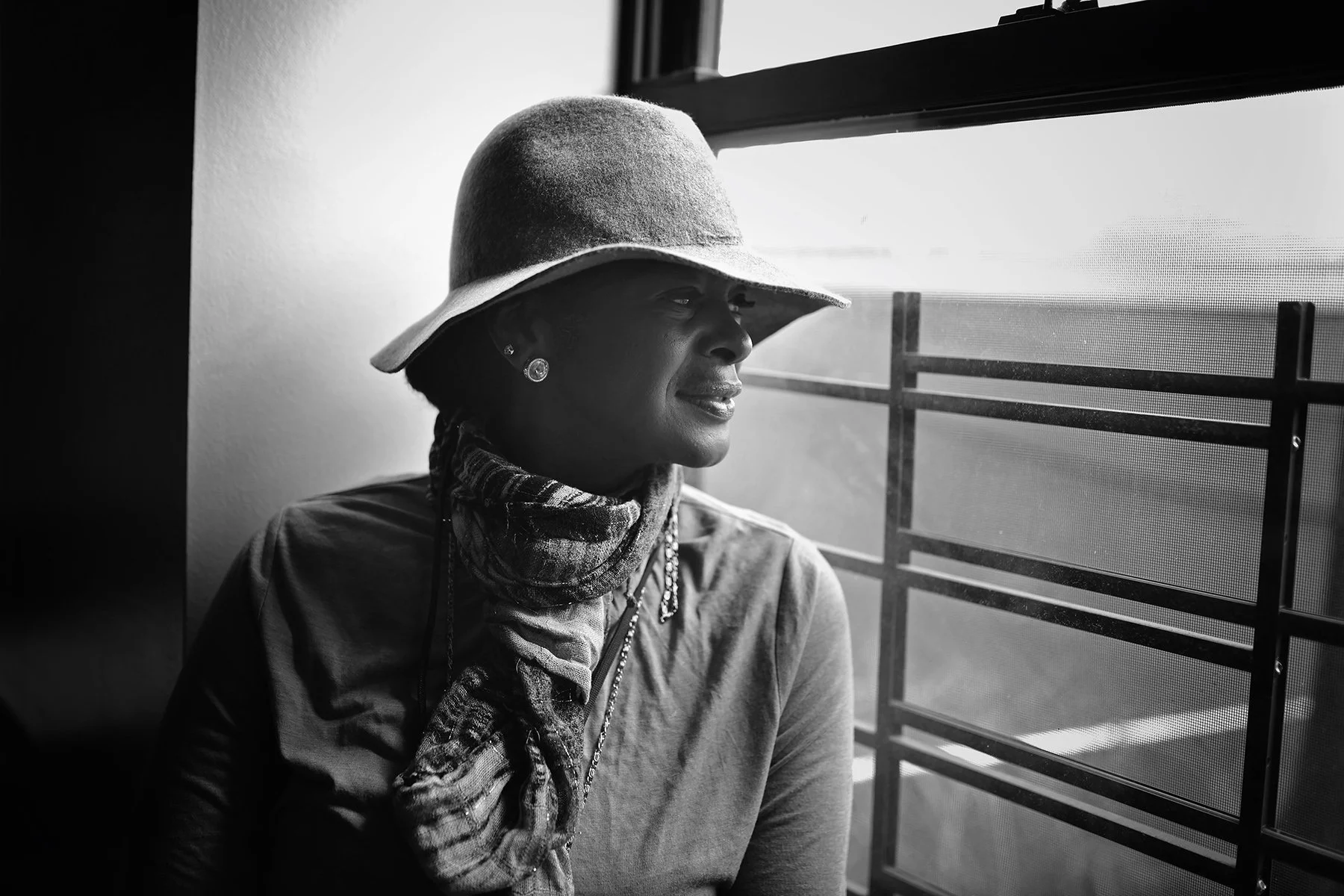 A woman wearing a large hat, earrings, and a scarf looks out a window on a sunny day, with metal bars and mesh screen visible outside.