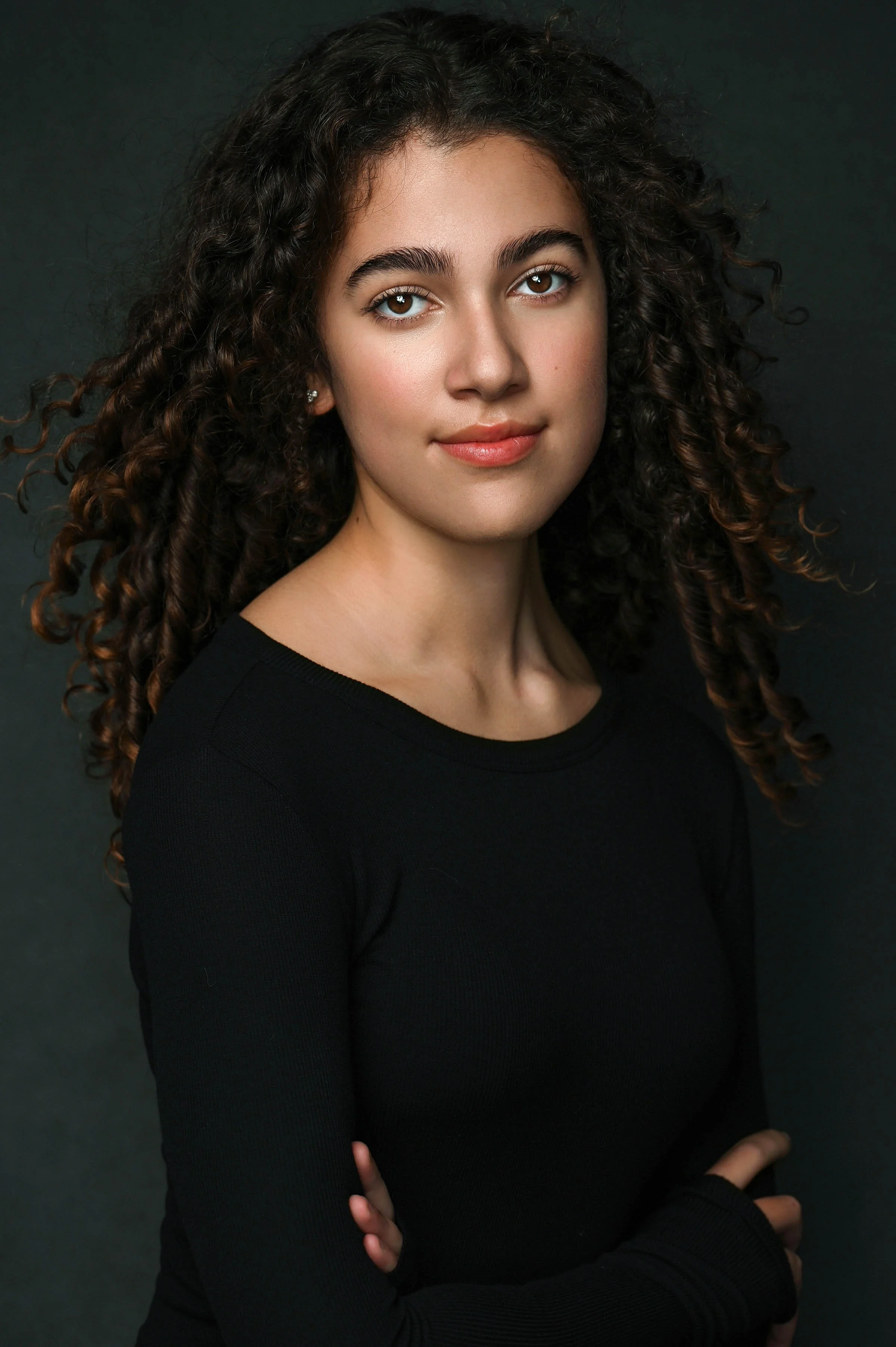A young woman with curly brown hair and earrings, wearing a black top, standing against a dark background.