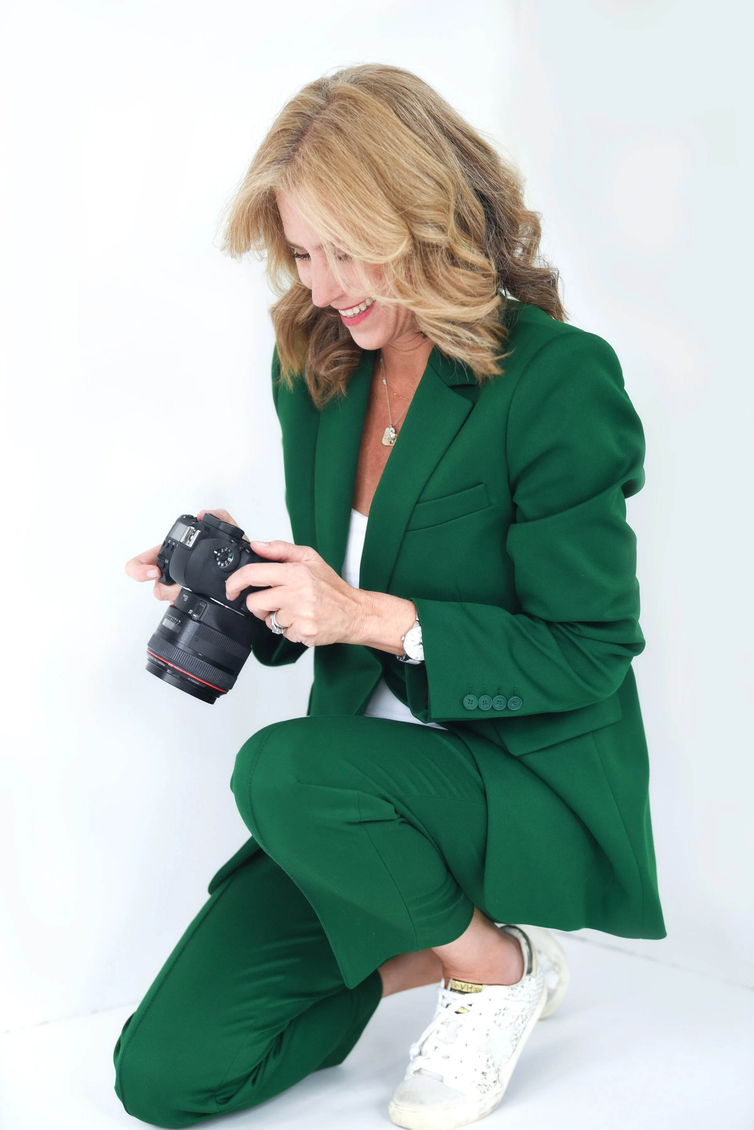 Woman with blonde and brown curly hair wearing a green suit, kneeling on one knee, holding a camera, smiling, against a plain white background.