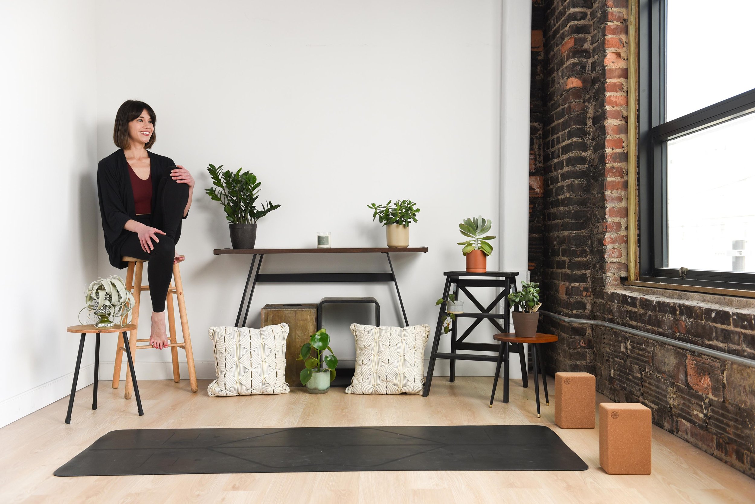 A woman practicing yoga or stretching in a bright, modern room with large windows, a white wall, and various potted plants on shelves and the floor, with a black yoga mat on the light wood floor.