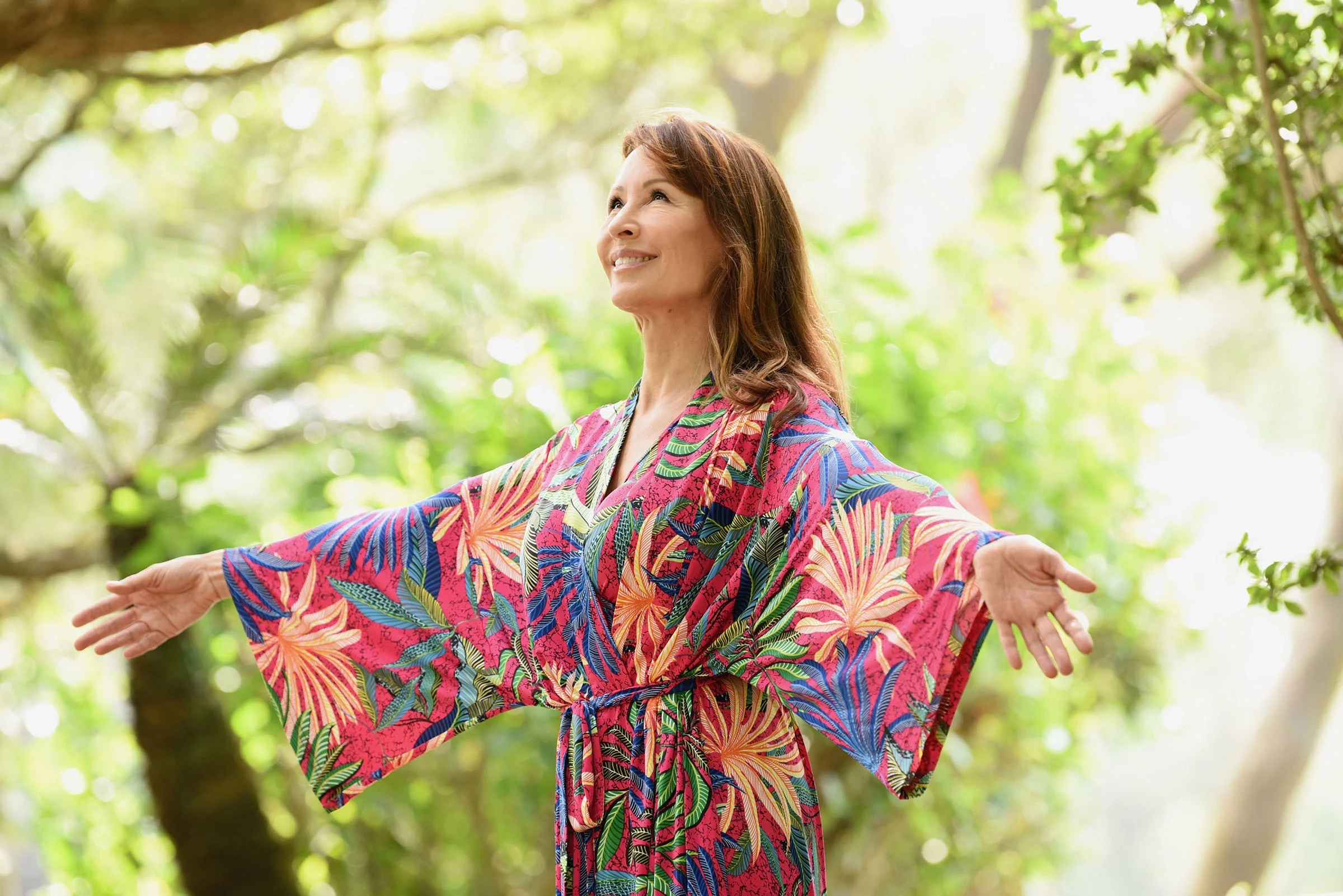 A woman in a colorful, tropical-patterned dress and with brown hair extending her arms outward while smiling and looking slightly upward in a lush, green outdoor setting with trees and bright dappled sunlight.