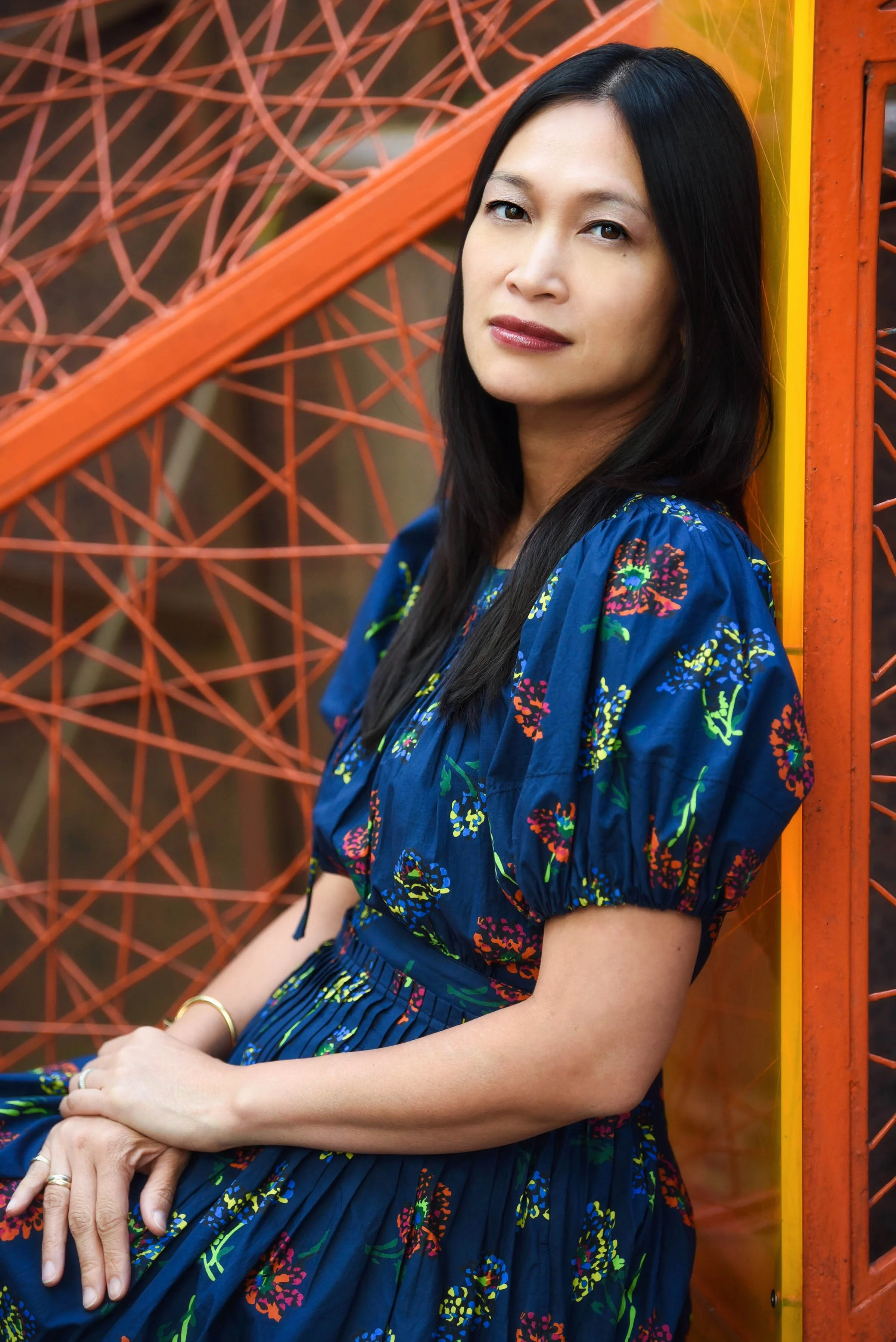 A woman with long black hair leaning against an orange metal structure with a web-like design, wearing a blue dress with a colorful floral pattern, and gold jewelry.