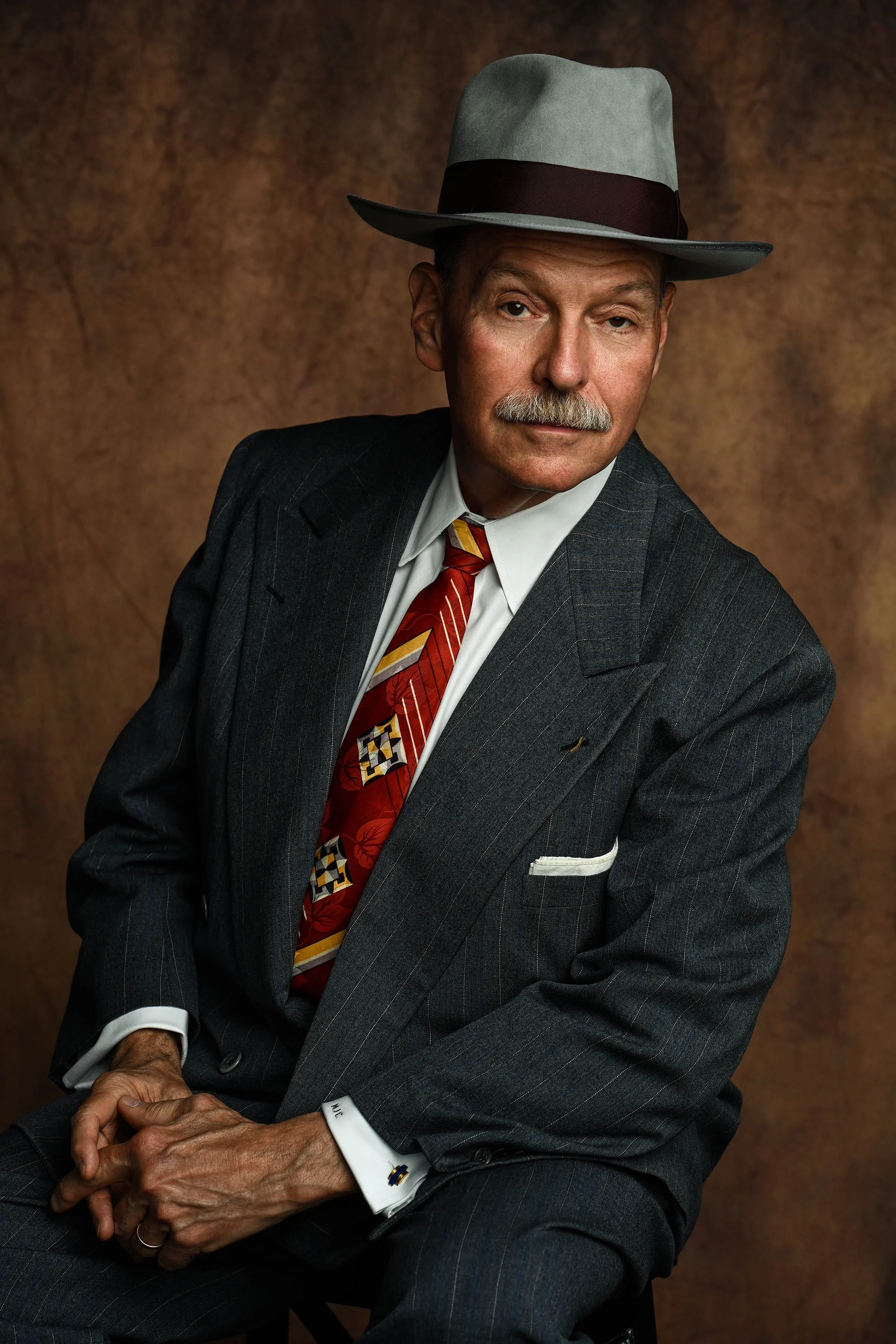 An elderly man with a mustache wearing a gray fedora hat, a dark pinstripe suit, a white dress shirt, and a colorful tie, sitting against a brown textured background.
