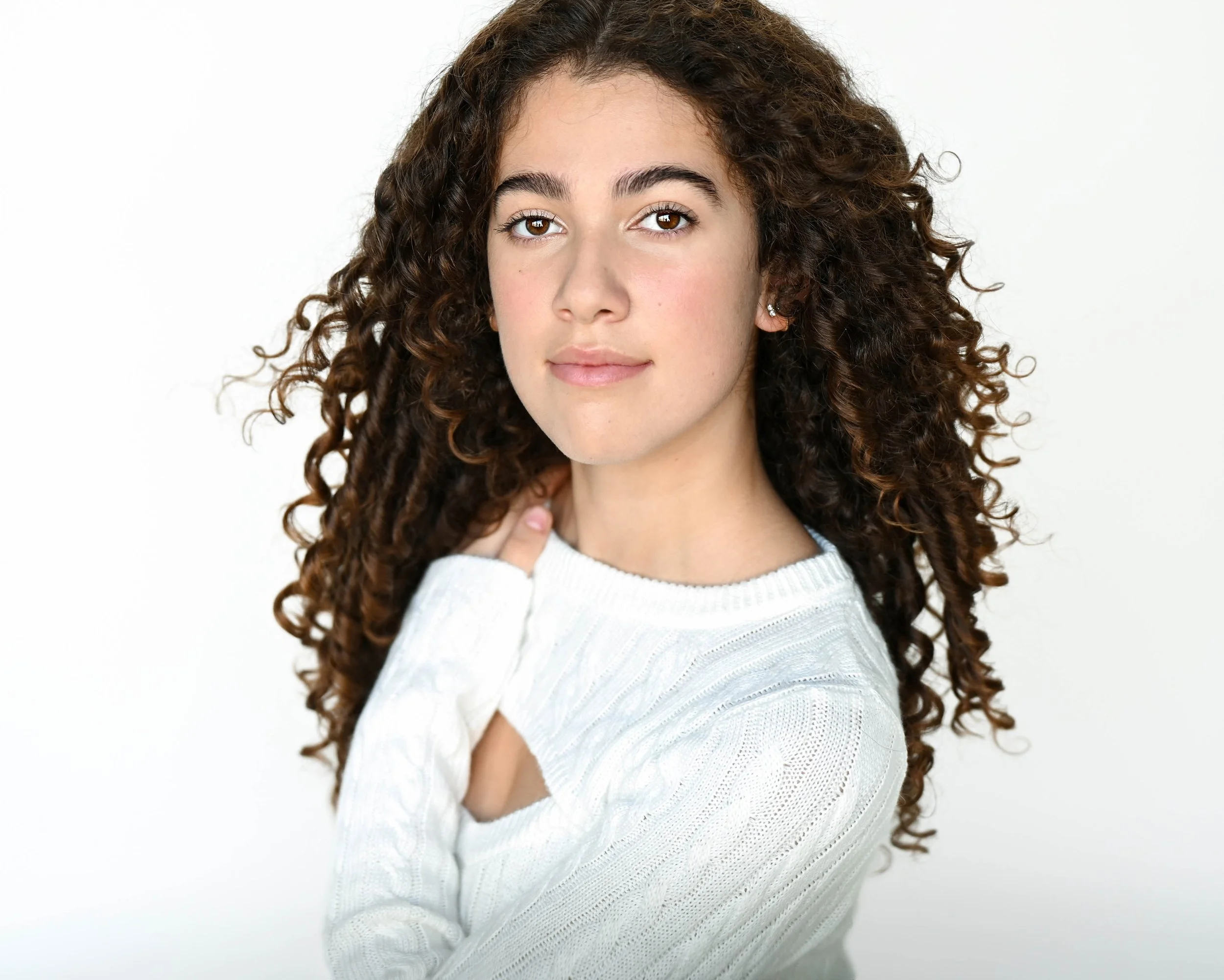 Portrait of a young woman with curly brown hair wearing a white knit sweater, standing against a plain white background.