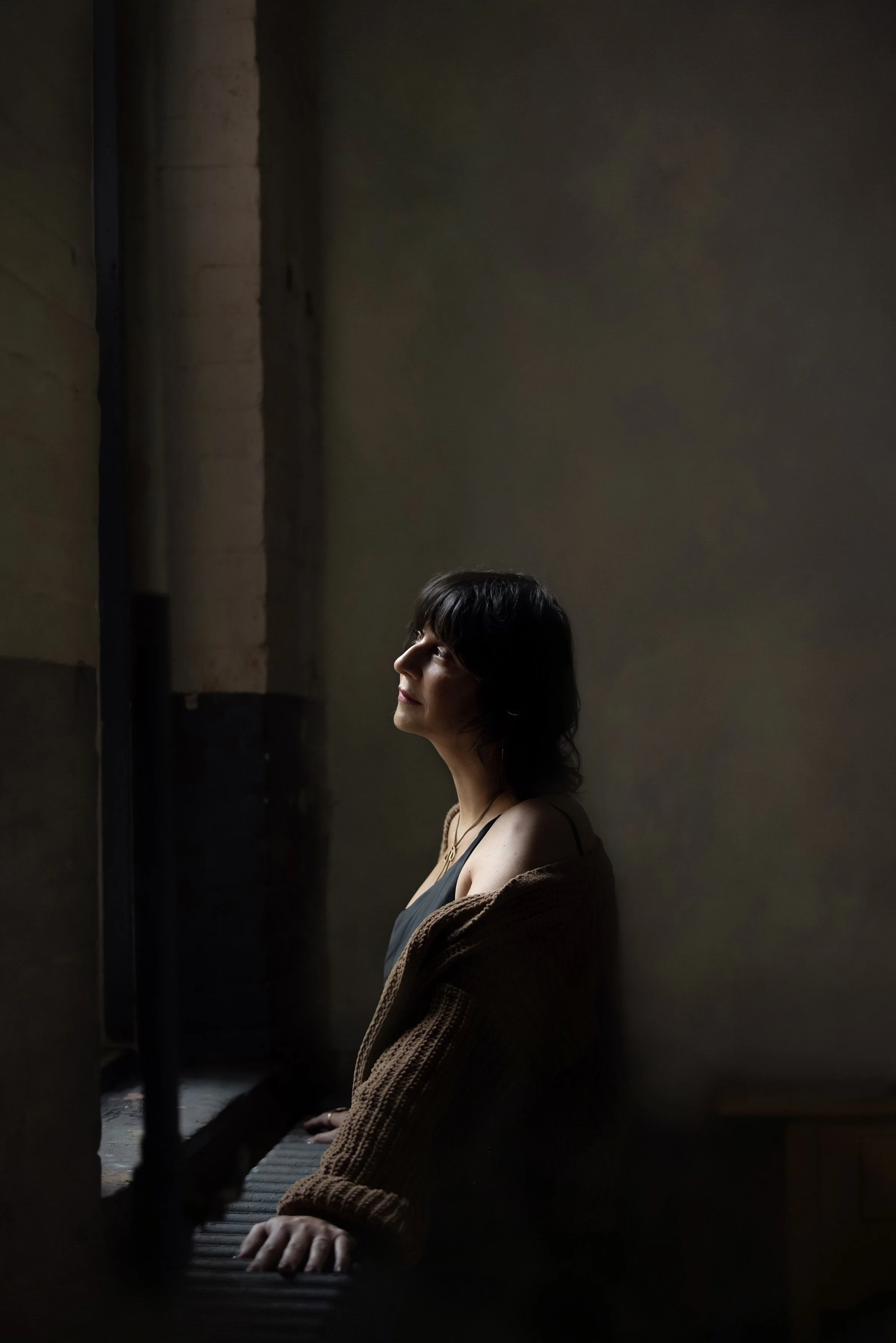 A woman sitting at a piano with her hands on the keys, wearing a brown jacket and a black top, in a dimly lit room.