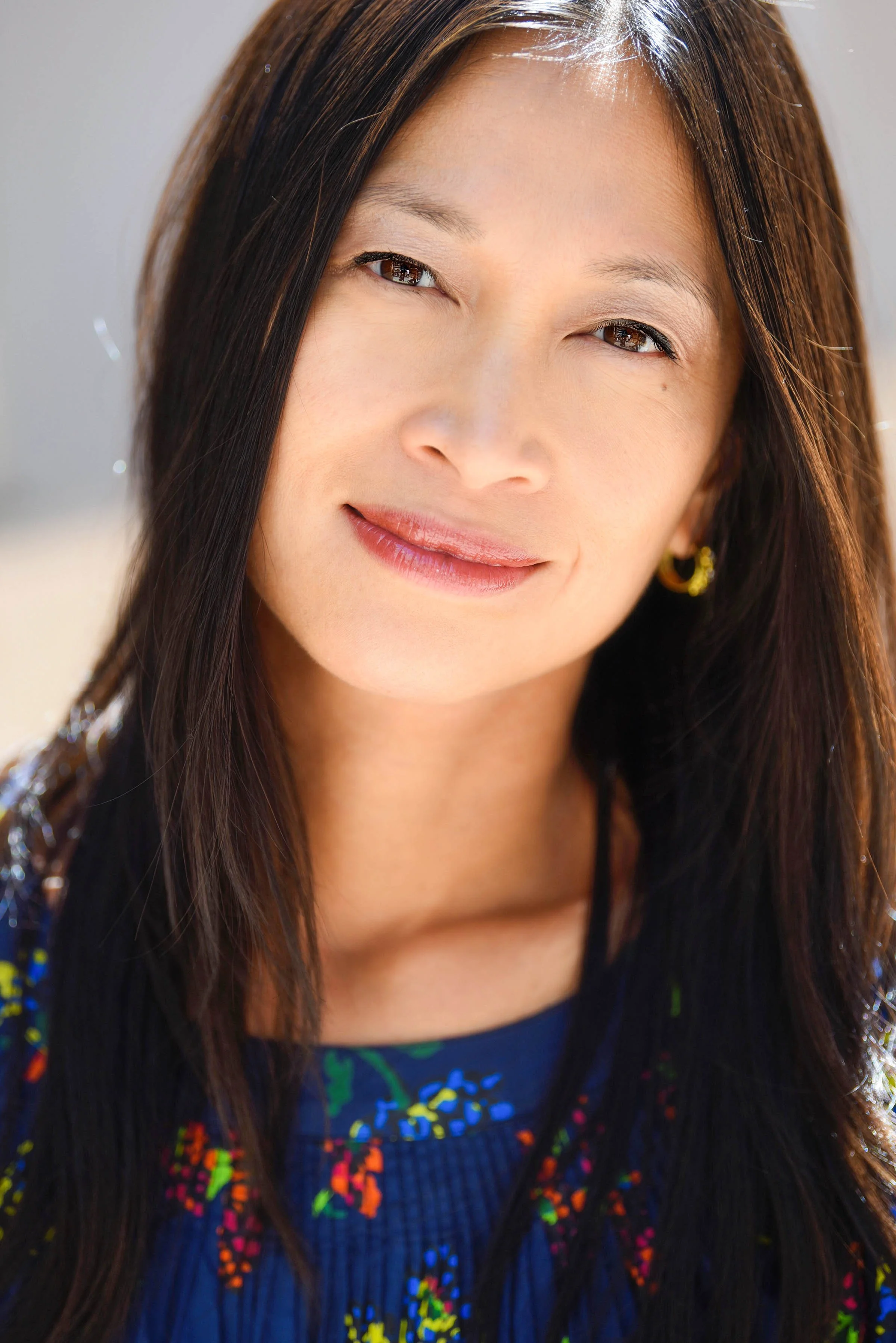 A woman with long, dark hair and light skin, smiling gently. She has light makeup and is wearing gold hoop earrings. She is dressed in a colorful, patterned top and a black open-front cardigan. The background is neutral and softly blurred.