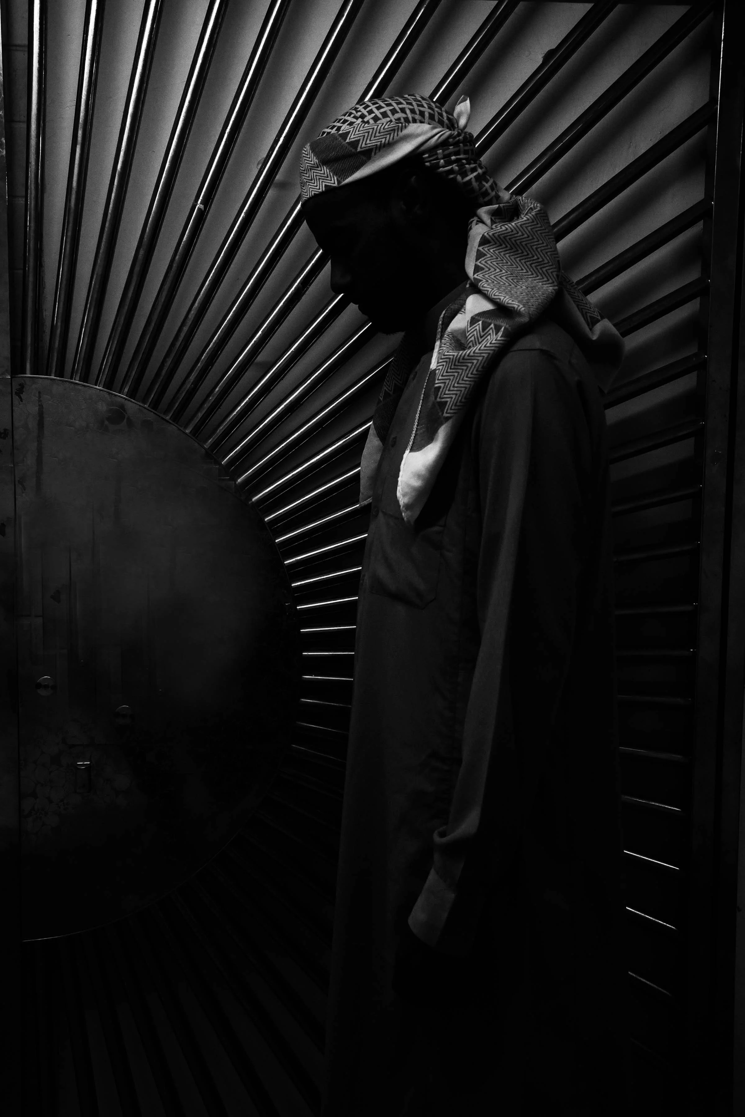 A silhouette of a person wearing a patterned headscarf and glasses, standing against a background of radial metal bars. The image is in black and white.
