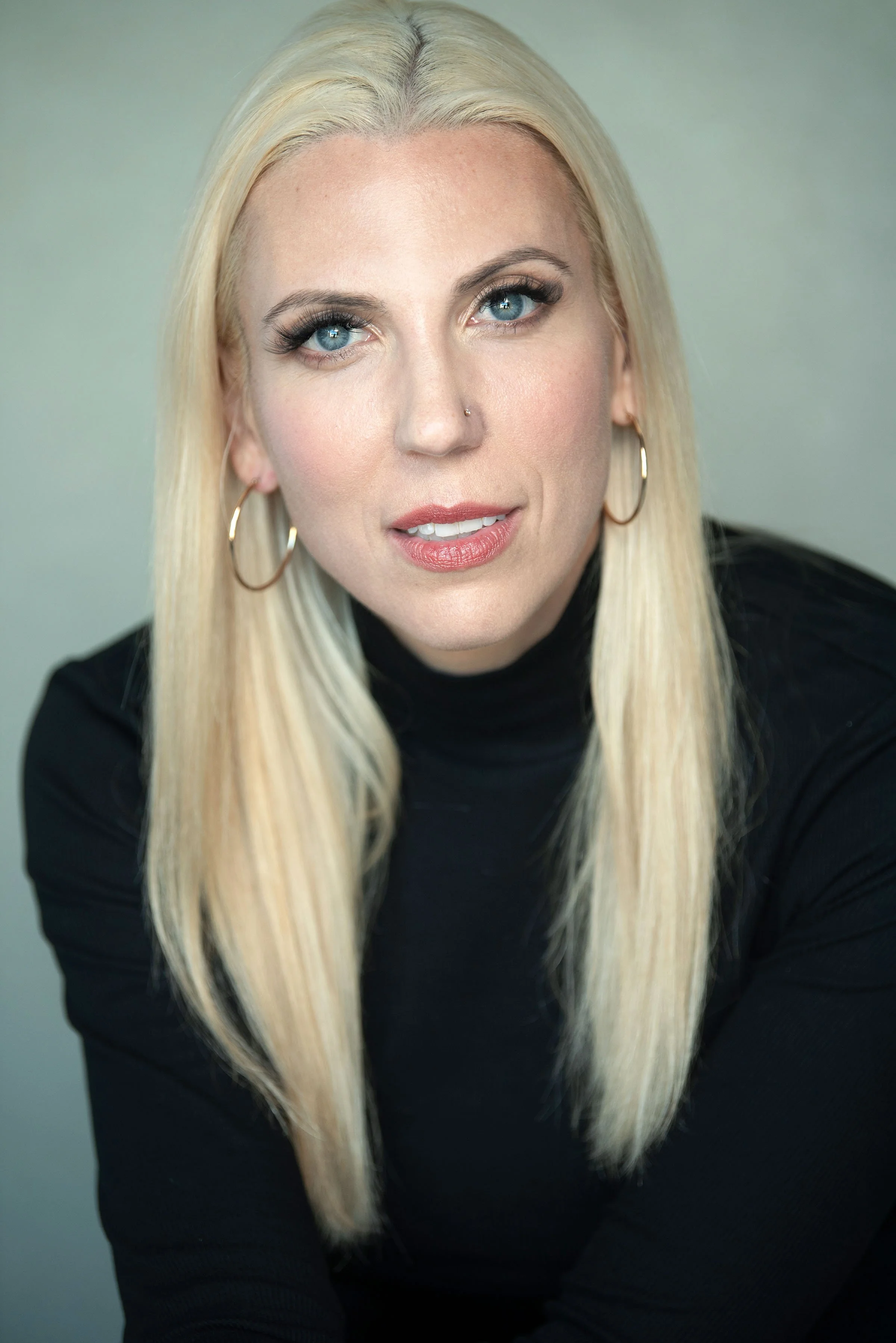 Close-up of a woman with long blonde hair, blue eyes, wearing a black top, hoop earrings, and makeup, looking at the camera.