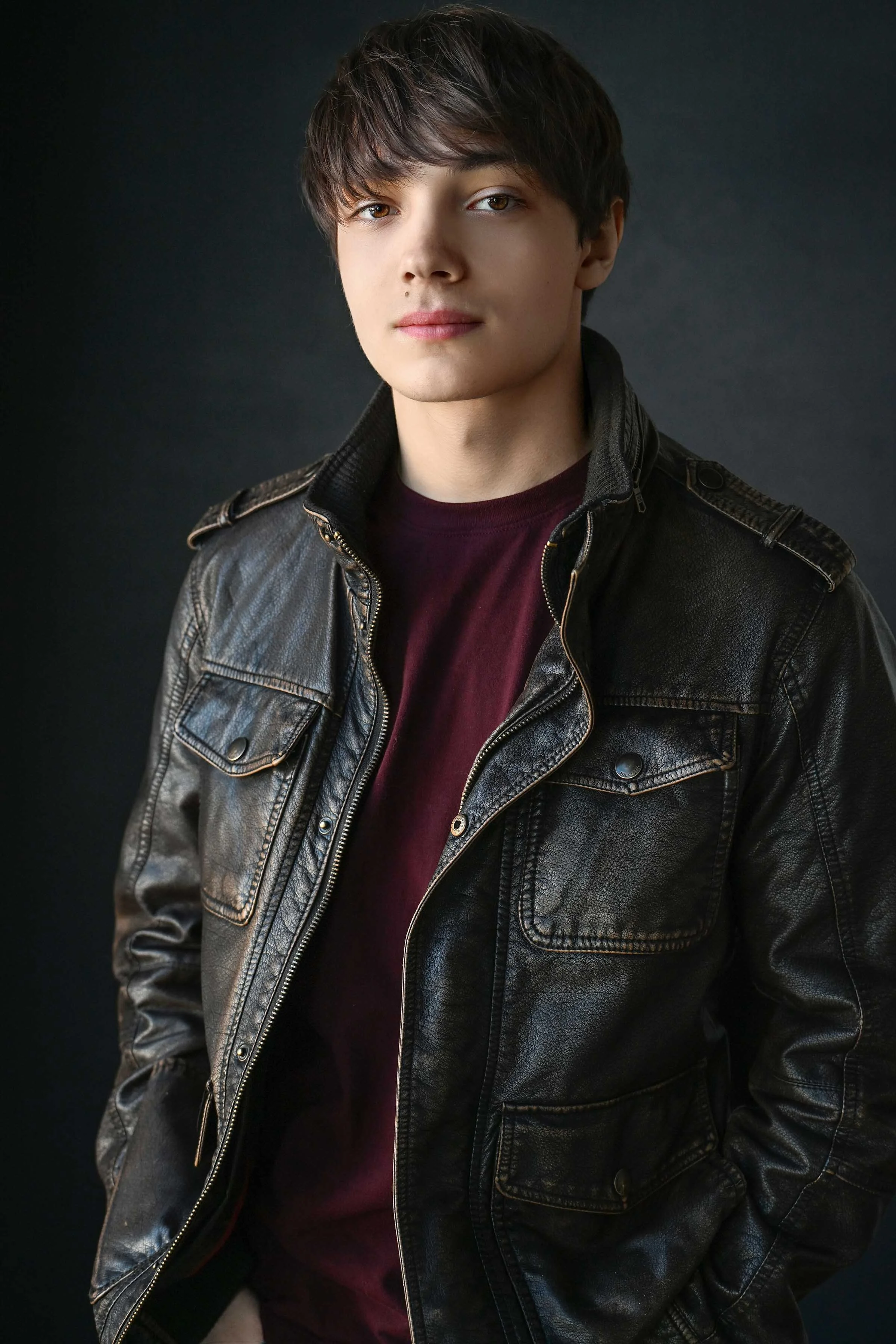 A young man with brown hair and fair skin wearing a black leather jacket over a maroon shirt, standing against a dark background.