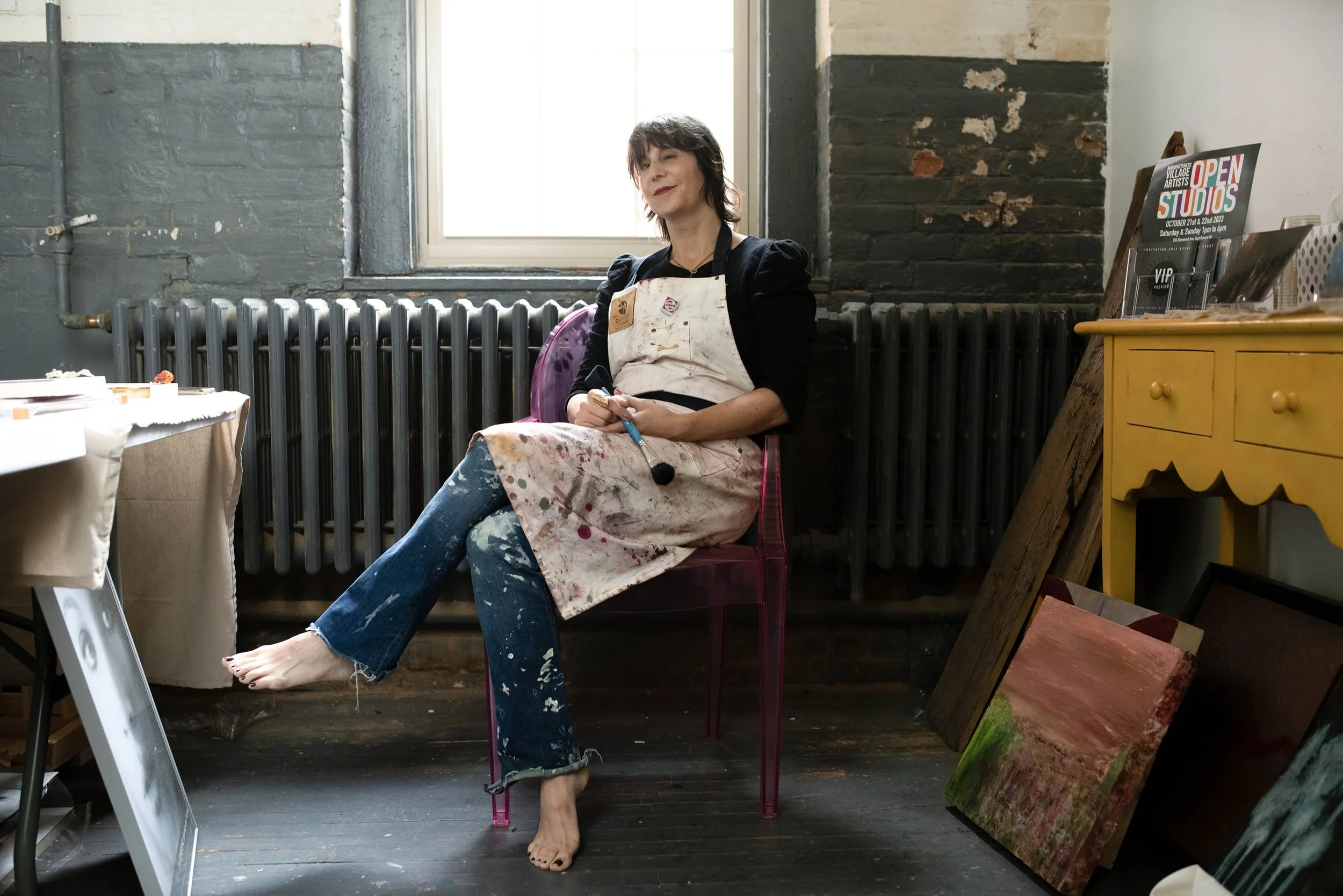 A woman with short hair, wearing a black top, jeans, and a worn apron, sits barefoot on a pink transparent chair in an artist's studio. She is holding a paintbrush and smiling softly. The studio has a large window, a yellow cabinet, some paintings le