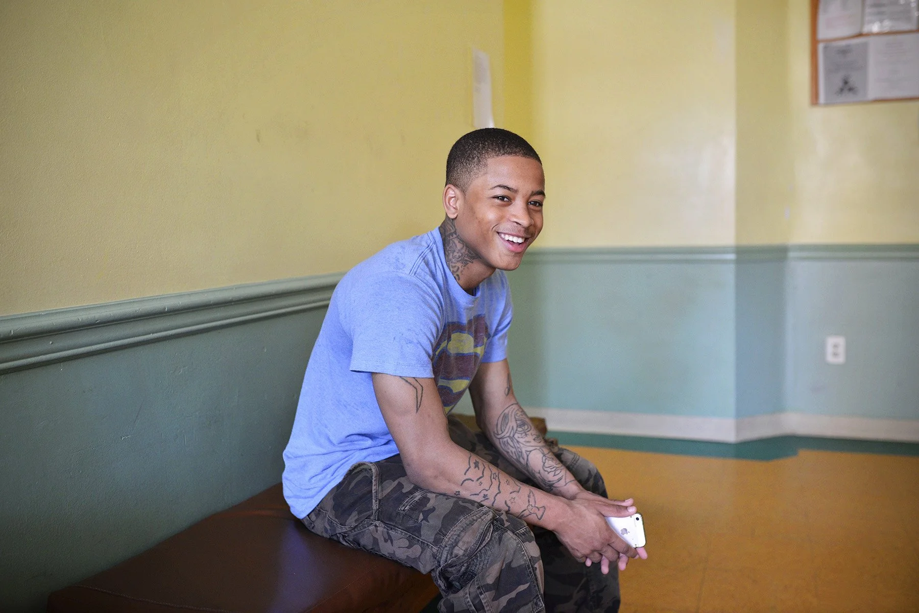 Young man with tattoos sitting on a bench in a waiting room, smiling, holding a smartphone.