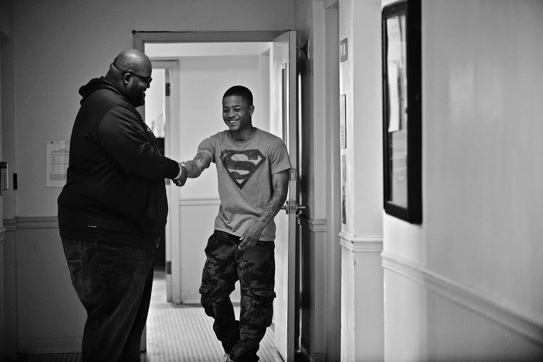 Two men, one with a bald head and glasses, and the other with short hair, tattoos, and a Superman t-shirt, shake hands in a hallway, smiling at each other.