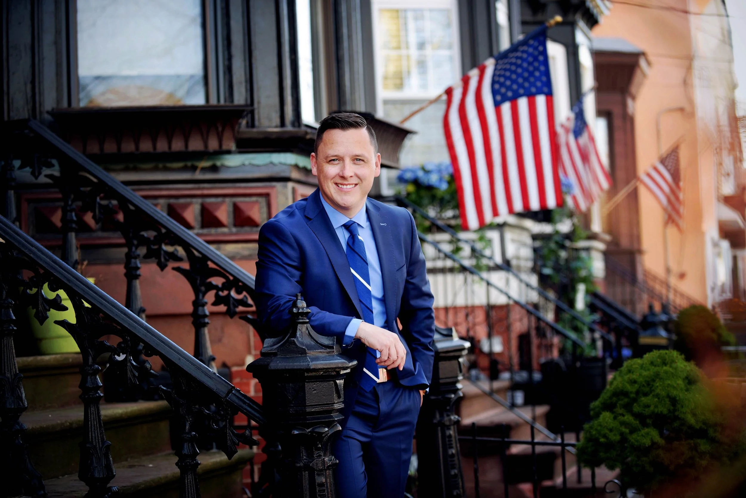 A man in a blue suit with a blue striped tie standing on the steps of a brownstone building, smiling at the camera, with American flags hanging in the background.