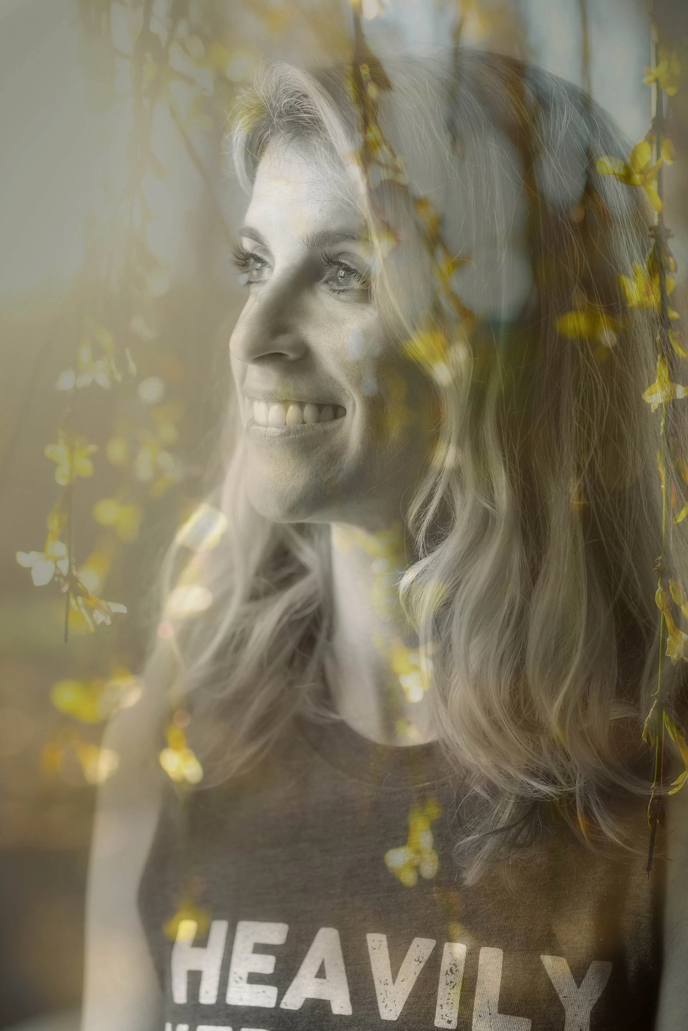 A woman with long, curly blonde hair smiling while looking to the side. There are yellow flowers and green leaves superimposed over her face. She is wearing a dark shirt with white letters spelling 'HEAVILY'.