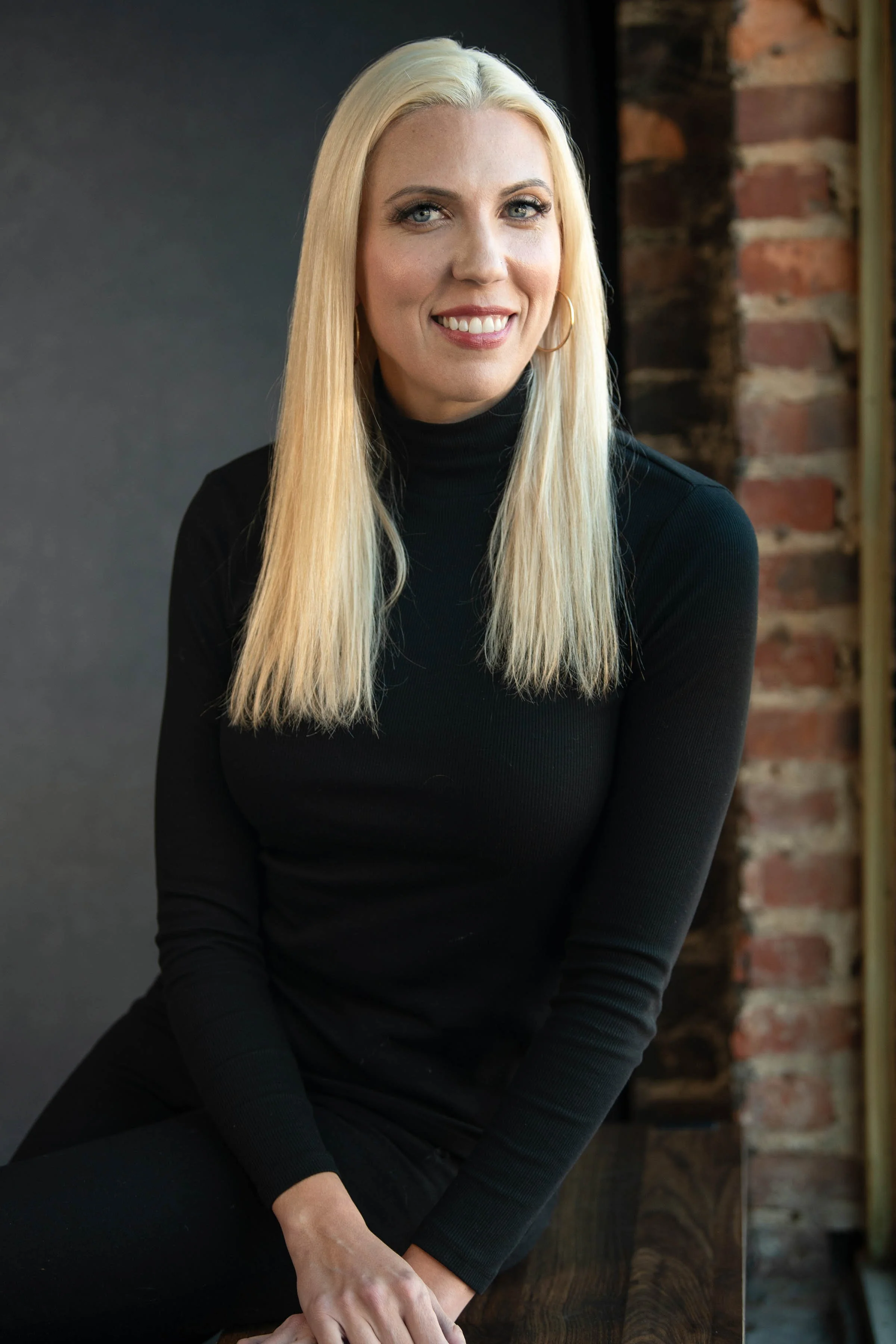 A woman with long blonde hair, blue eyes, and hoop earrings smiling, wearing a black turtleneck, sitting at a wooden table with a brick wall in the background.