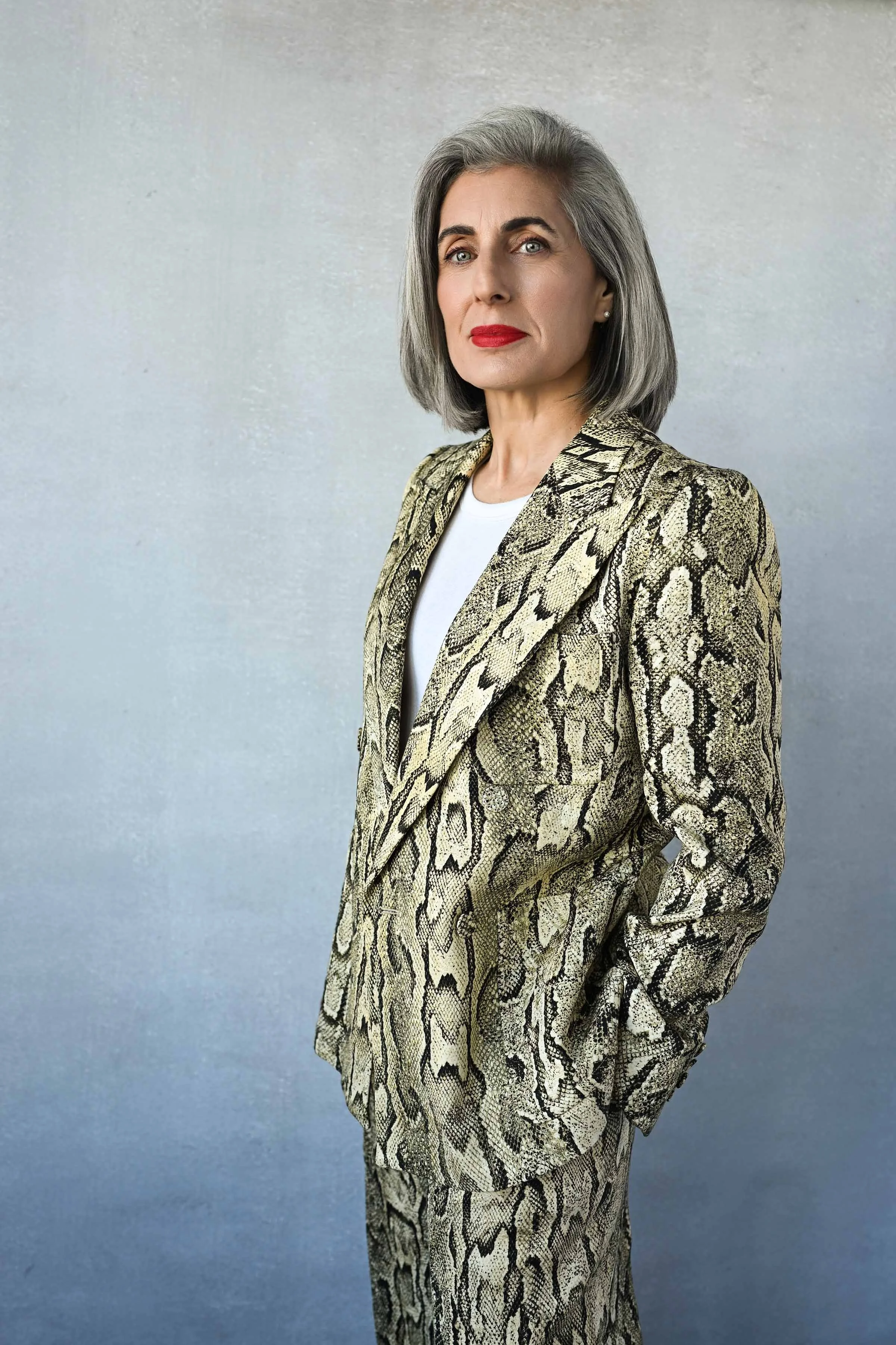 A woman with shoulder-length gray hair, wearing a snakeskin-patterned blazer and matching pants, posing against a plain gray background.