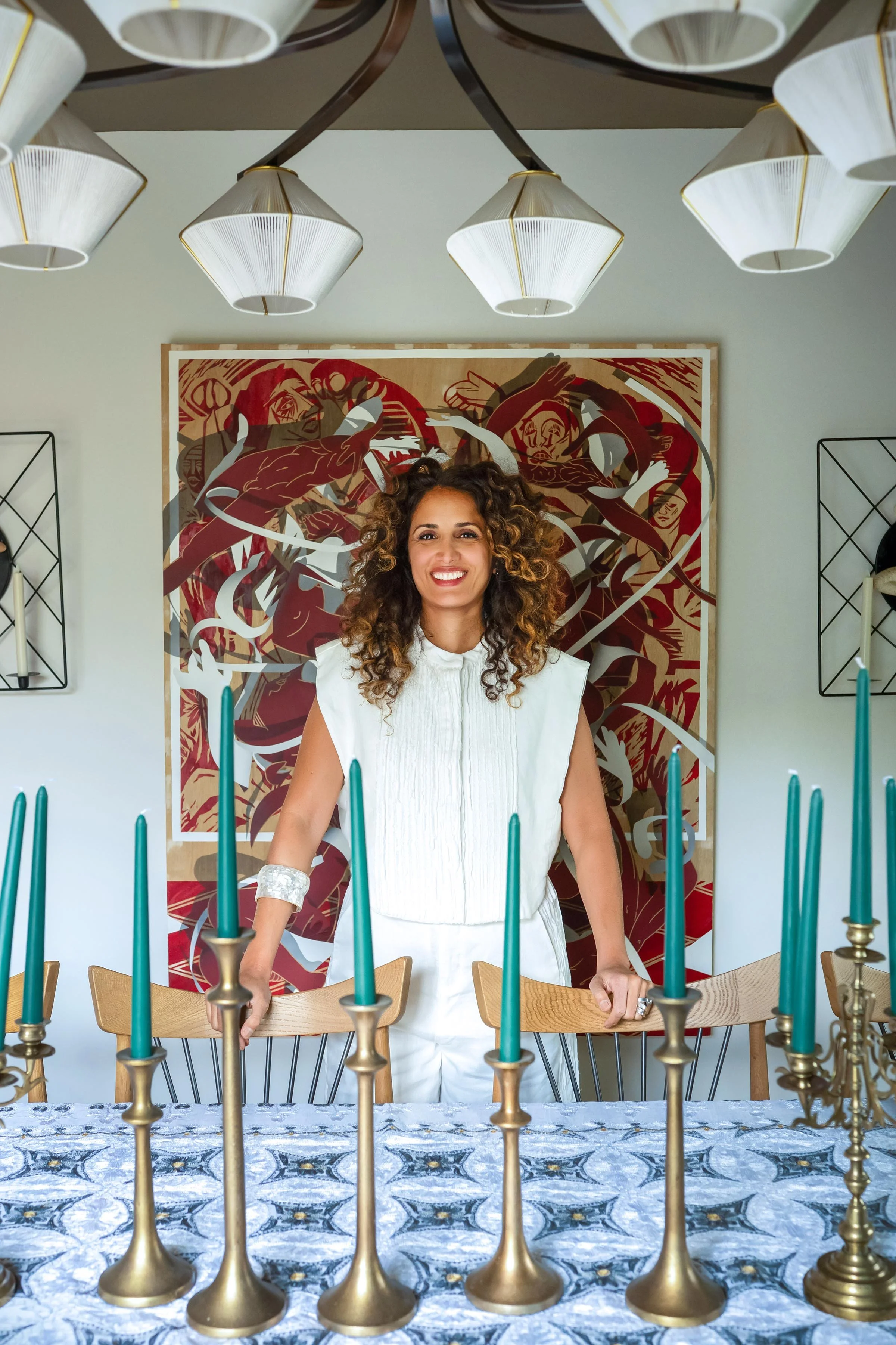 A woman with curly hair standing behind a dining table with blue candles in brass candle holders, in front of a large abstract artwork.