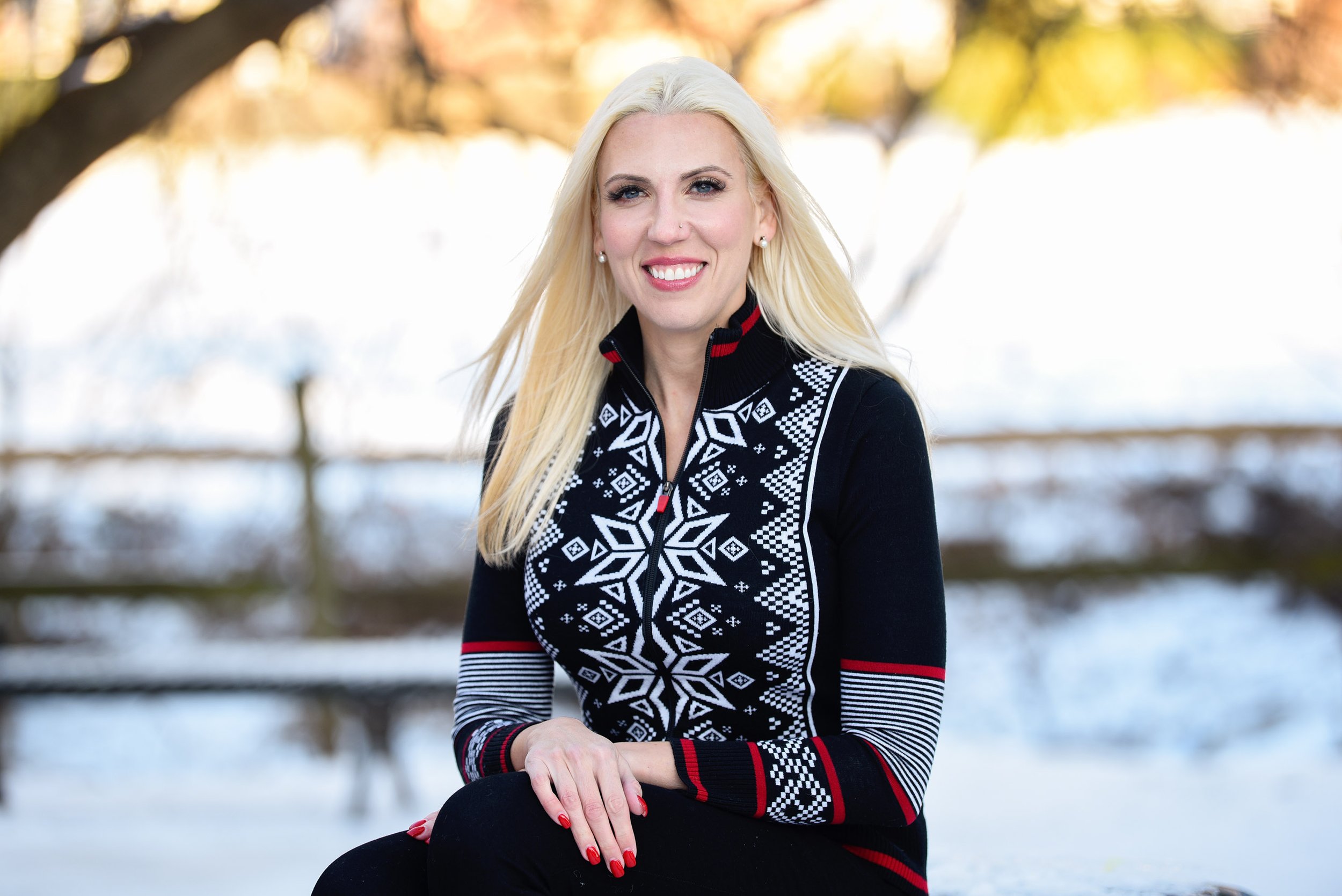 A woman with long blonde hair smiling outdoors in winter, wearing a black and white patterned sweater with red accents, and black pants, with snow and trees in the background.