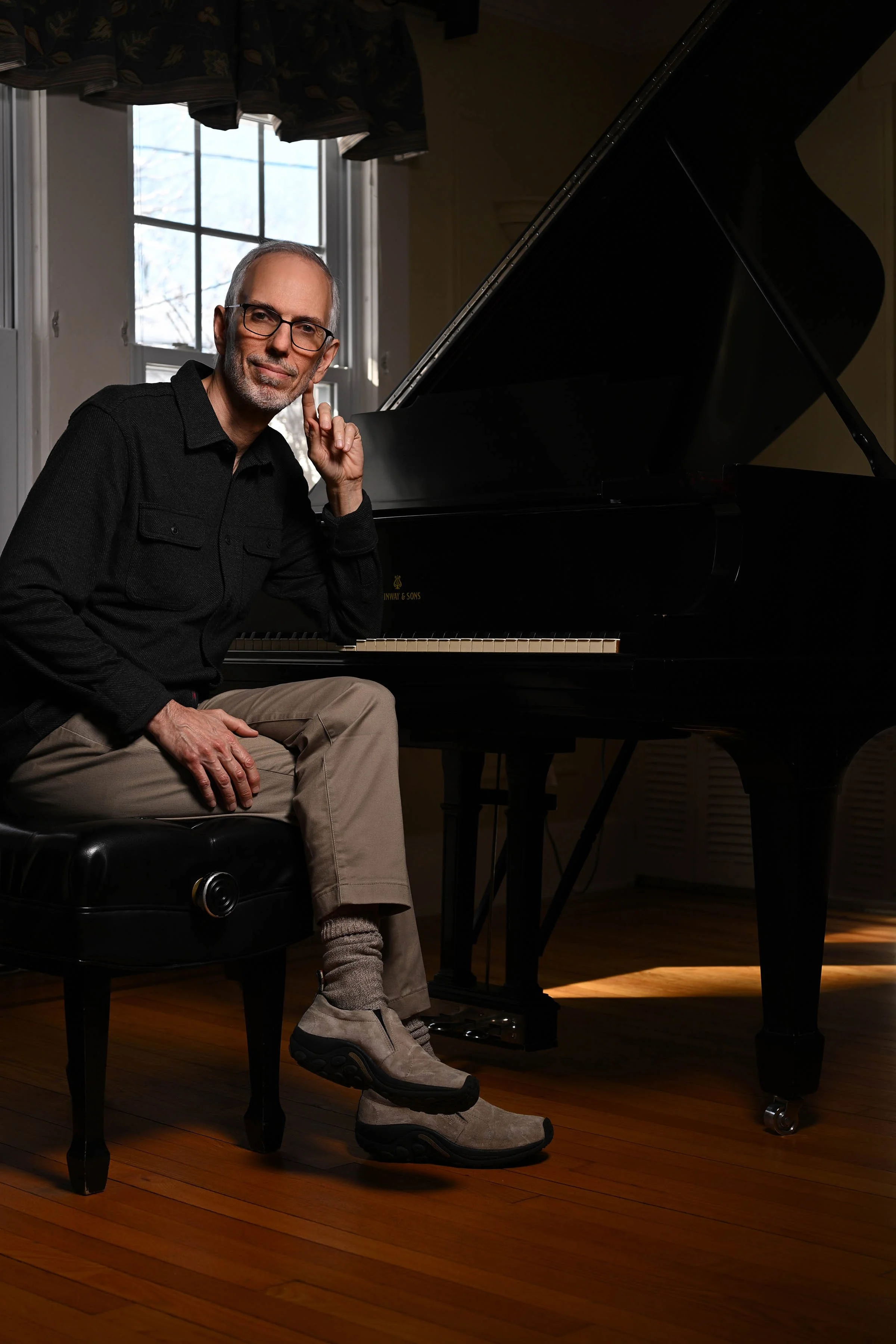 A man with glasses and a beard sitting on a black piano bench in a room with hardwood floors, next to a black grand piano, with a large window in the background.