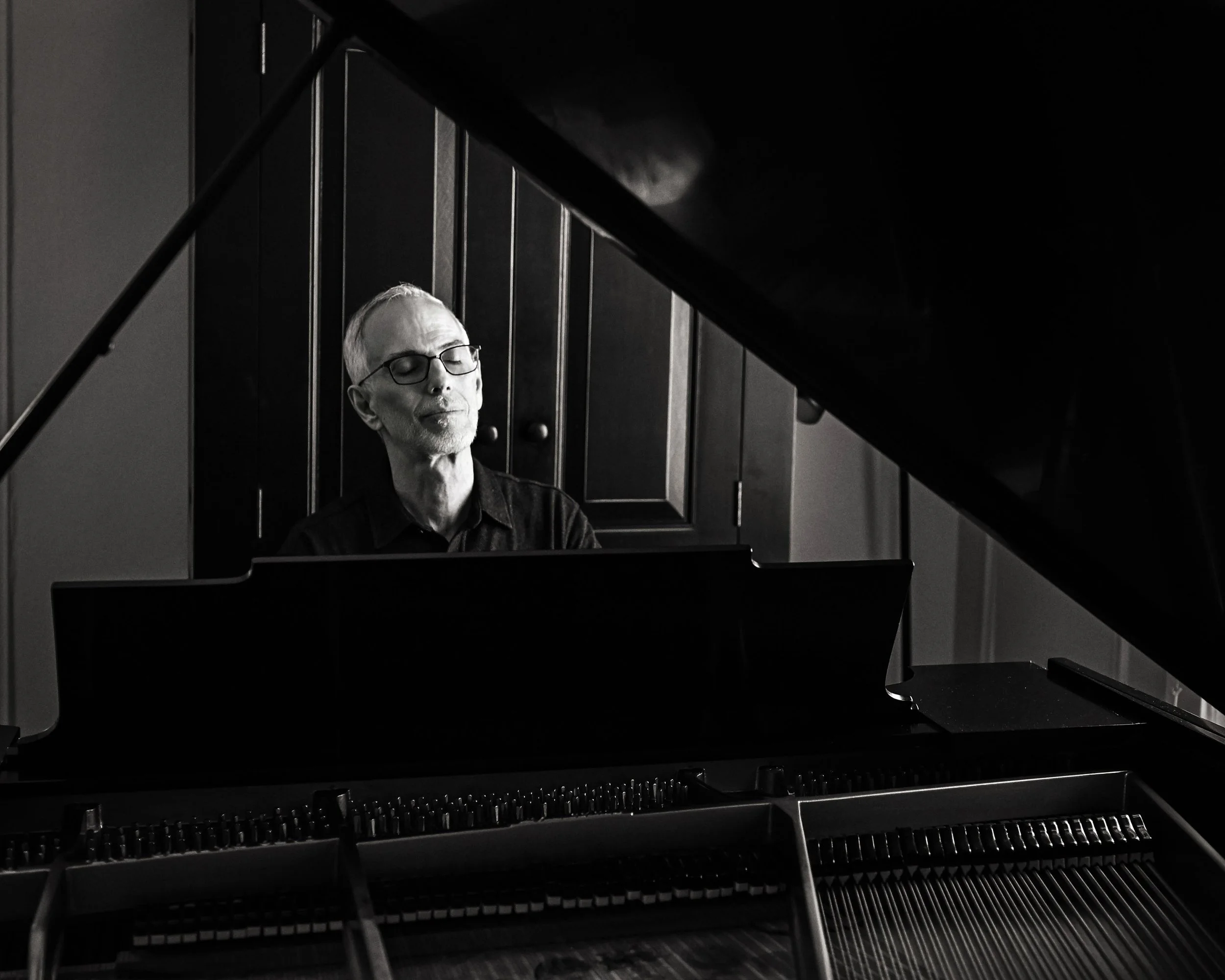 A person with glasses playing a grand piano, reflected in the piano's glossy black surface.