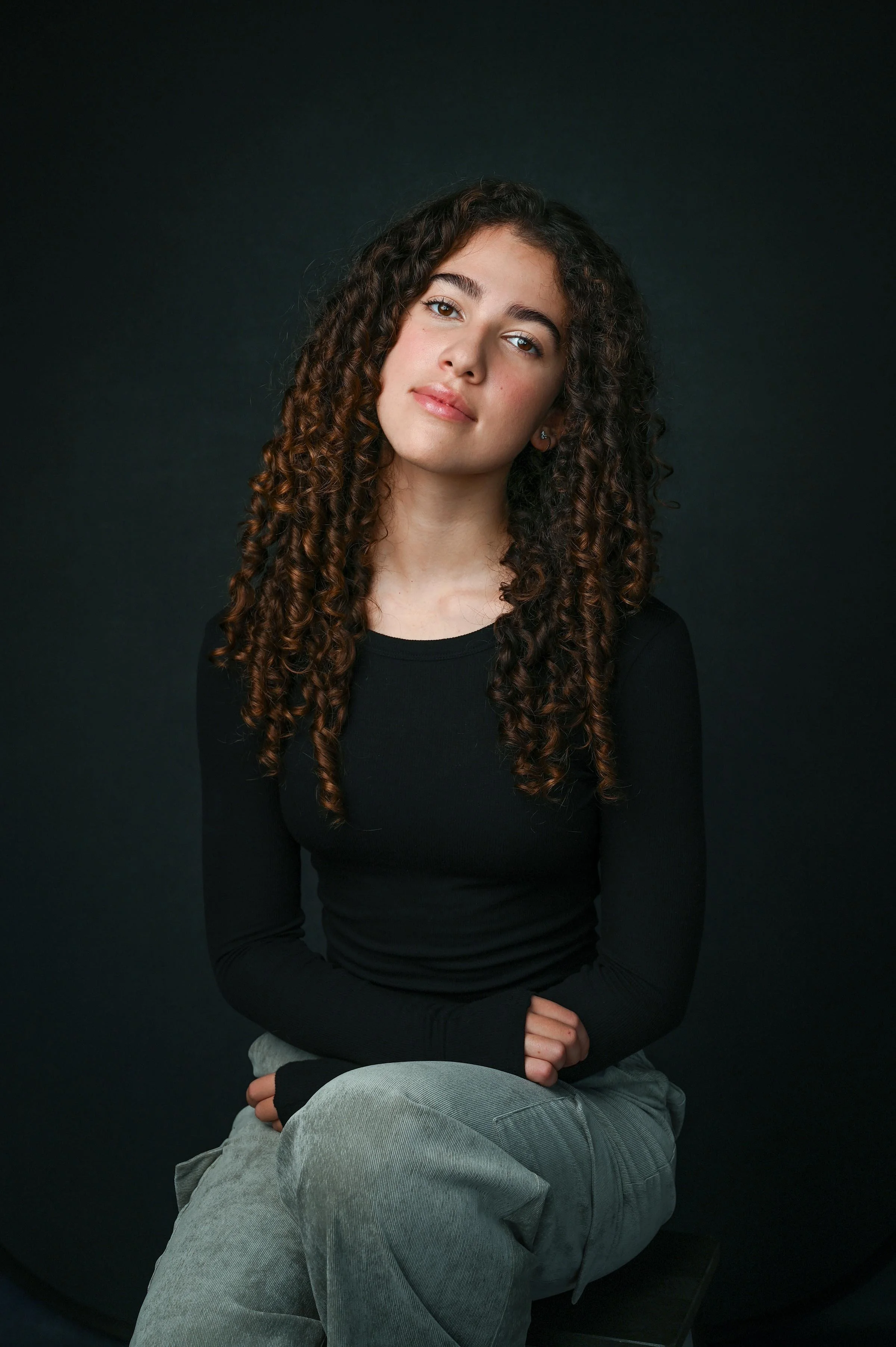 A young woman with long, dark, curly hair and light skin sitting against a dark background. She is wearing a black long-sleeve shirt and gray pants, with her hands resting on her lap. She has a calm expression and looks directly at the camera.