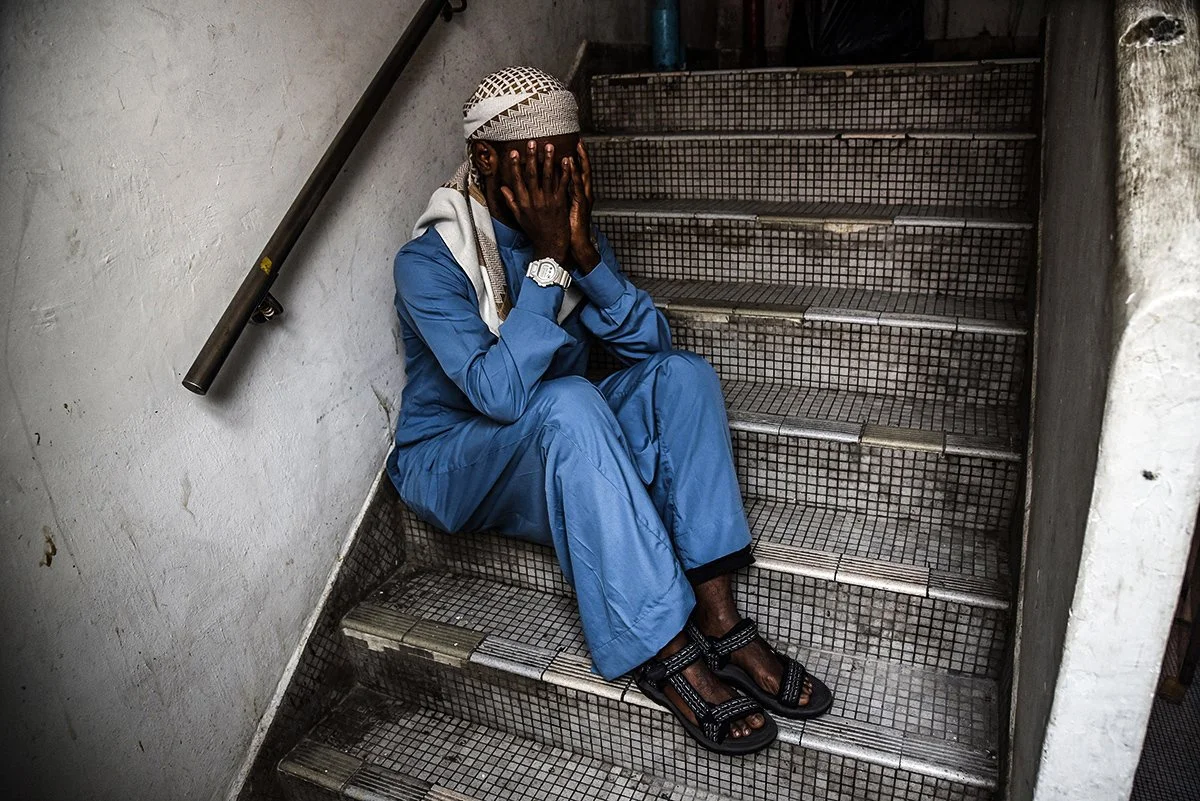 A man wearing a blue jumpsuit and sandals sitting on stairs with his hands covering his face.