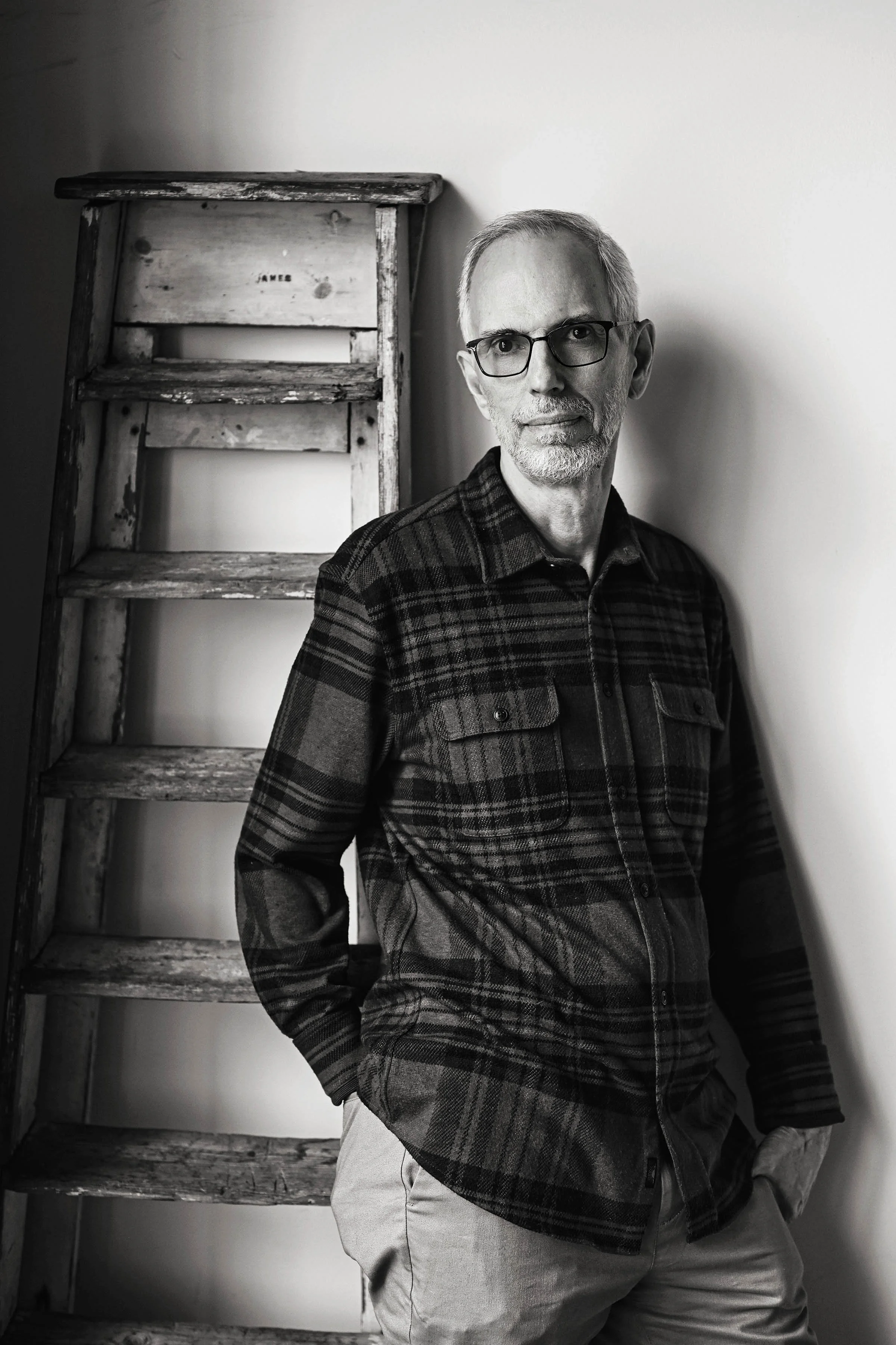 A black and white photo of an older man with glasses, a gray beard, and short gray hair, wearing a plaid shirt and beige pants. He is standing in front of a plain wall next to a weathered wooden ladder with the name "James" visible on one of its step
