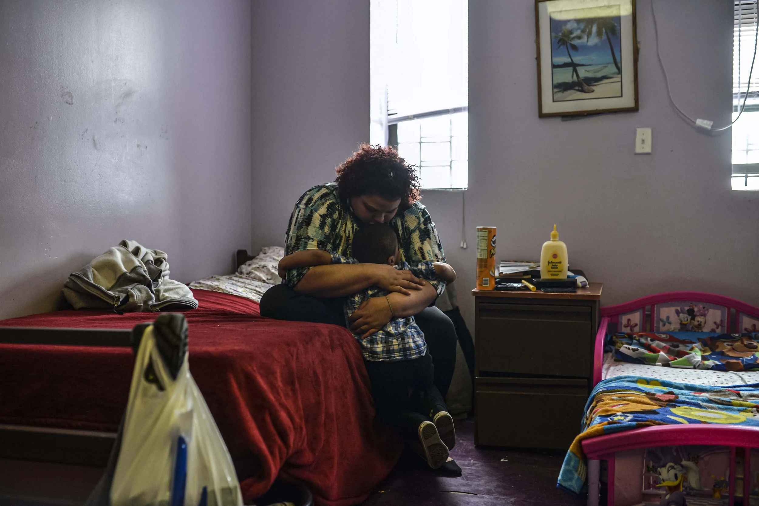 A woman sitting on a bed hugging a young child in a bedroom with plain walls, small window, and various household items.