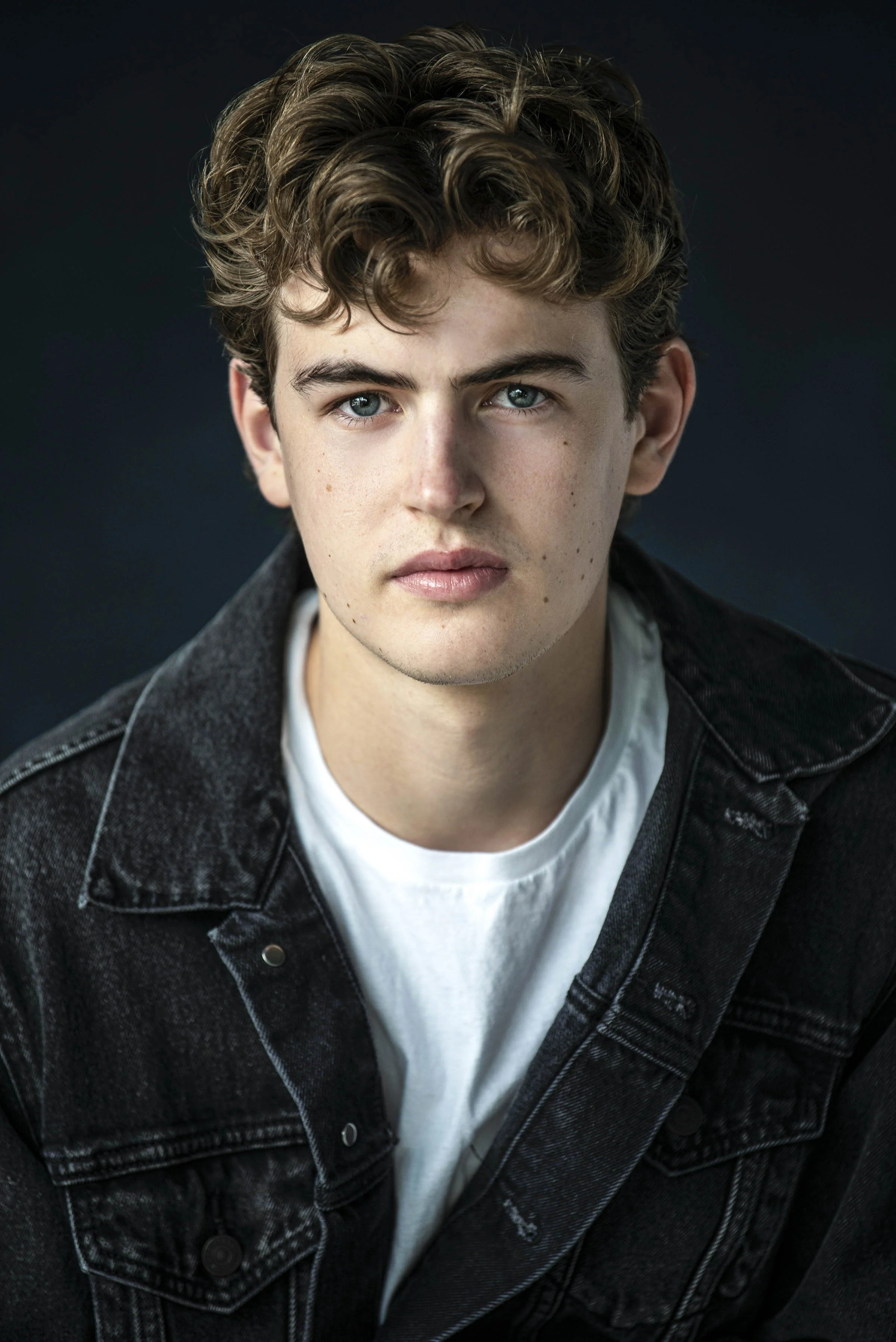 Close-up of a young man with curly brown hair, blue eyes, wearing a white T-shirt and a black denim jacket, against a dark background.