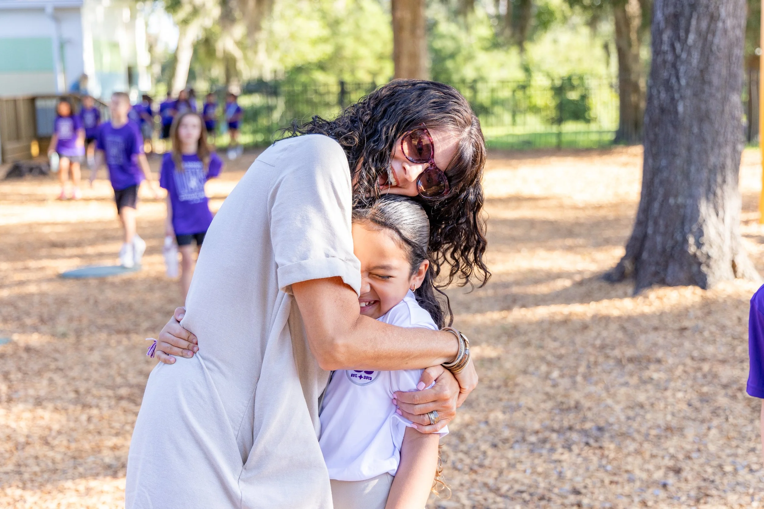 A woman with curly hair and sunglasses hugging a young girl, both smiling, outdoors in a park with trees and children in purple shirts in the background.