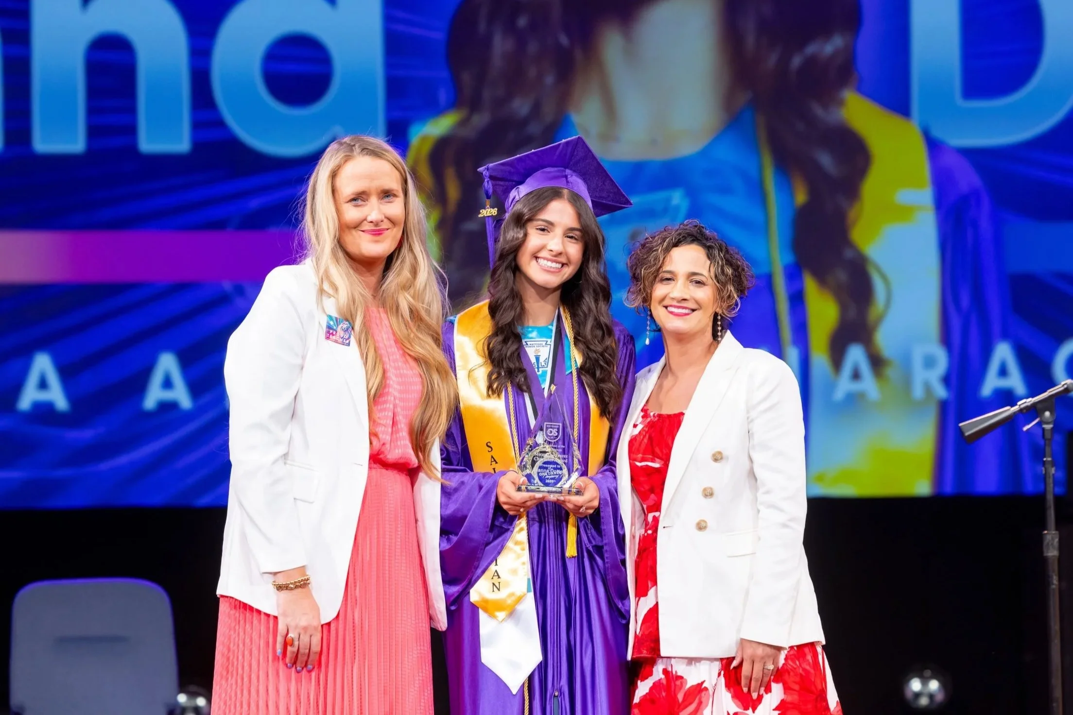 A graduation ceremony with a young female graduate in a purple cap and gown holding a trophy, flanked by two women in formal attire, on stage with a colorful backdrop.