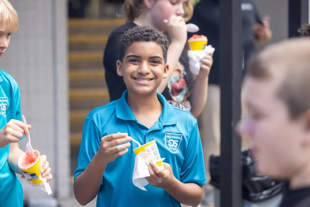 Smiling boy in blue school uniform holding a yellow cup of ice cream with a spoon at a school event.