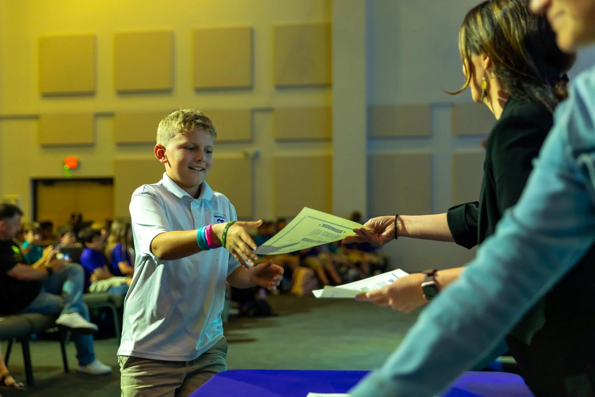 A young boy in a white shirt receiving a certificate from a woman at an indoor event with many people seated in the background.