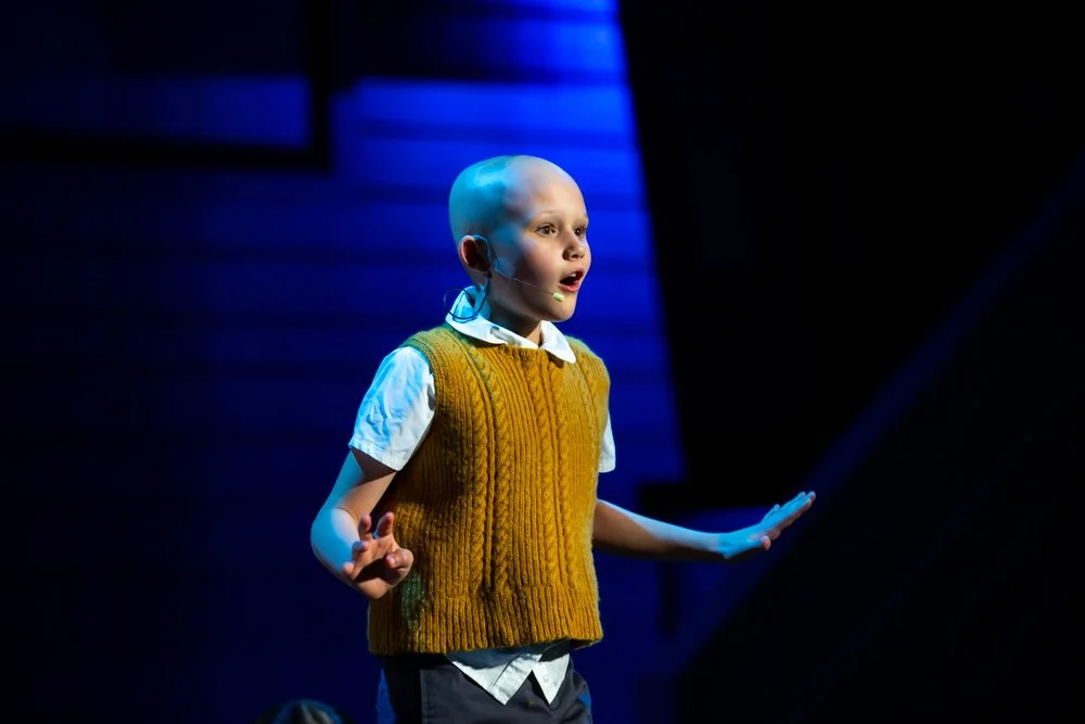 Child actor with bald head and makeup, wearing a mustard sweater vest over a white shirt, speaking into a microphone headset on a stage with dark and blue background.