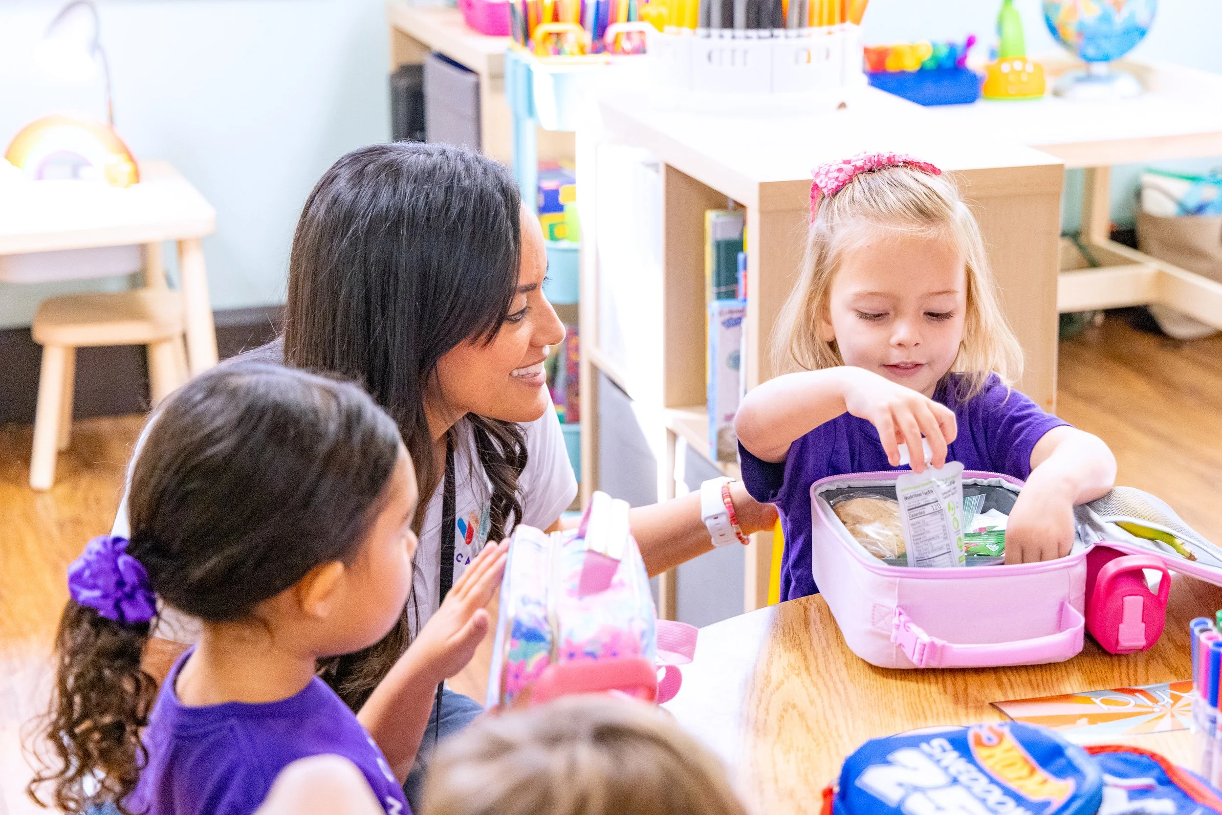 A group of young girls and a woman with dark hair sitting at a wooden table in a colorful classroom, with a girl opening her lunchbox.