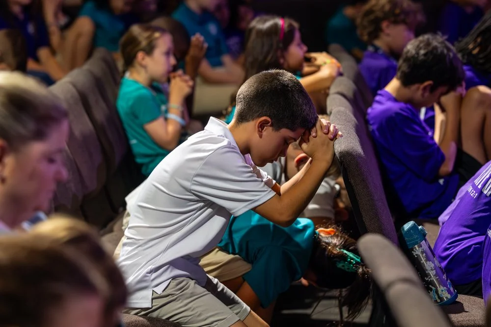 A group of children sitting in a church or auditorium, with one boy in the foreground praying with his head bowed and hands clasped.