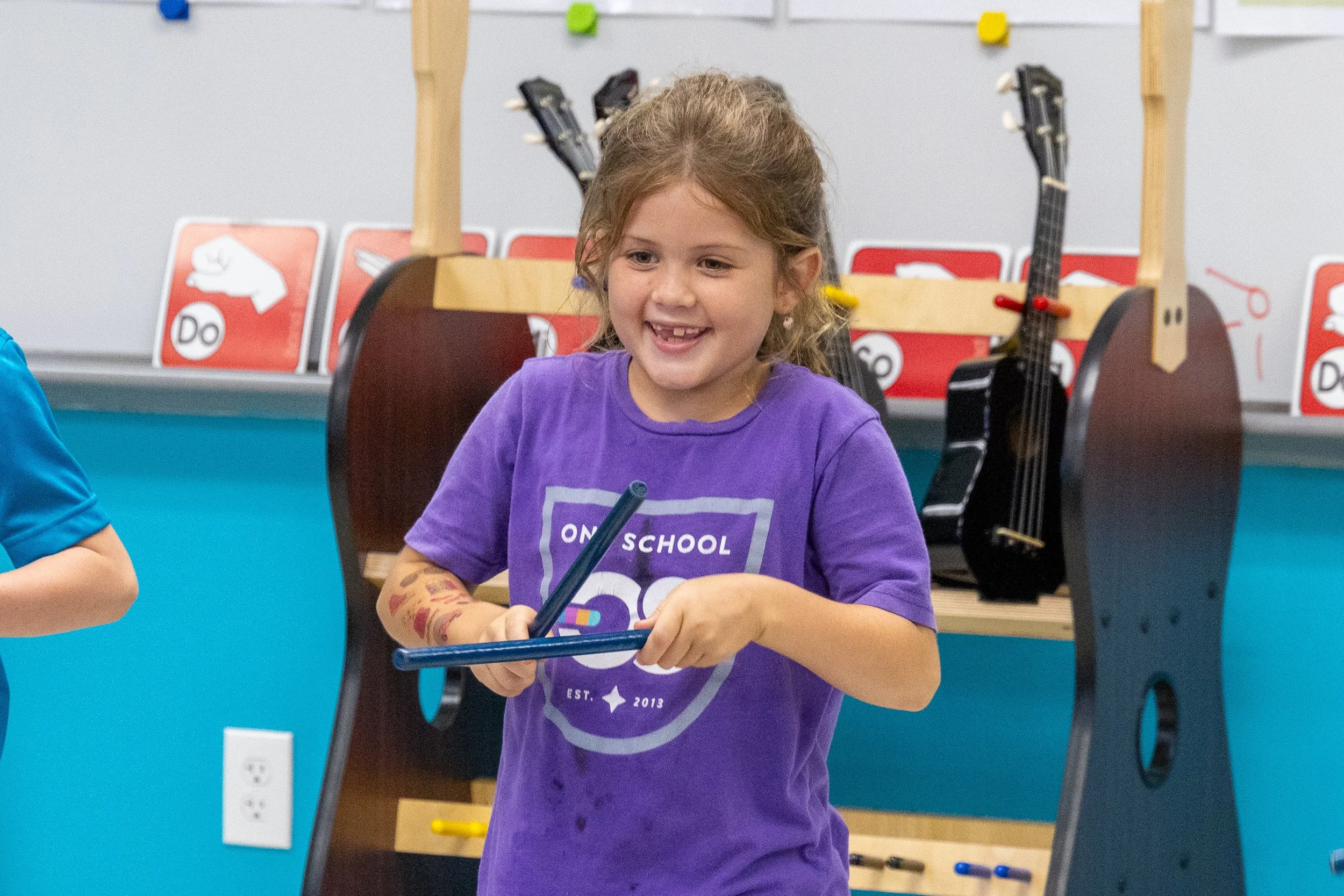 A young girl with curly hair wearing a purple T-shirt with a logo, smiling and holding a tablet in her hands, in a classroom setting with musical instruments and educational posters in the background.