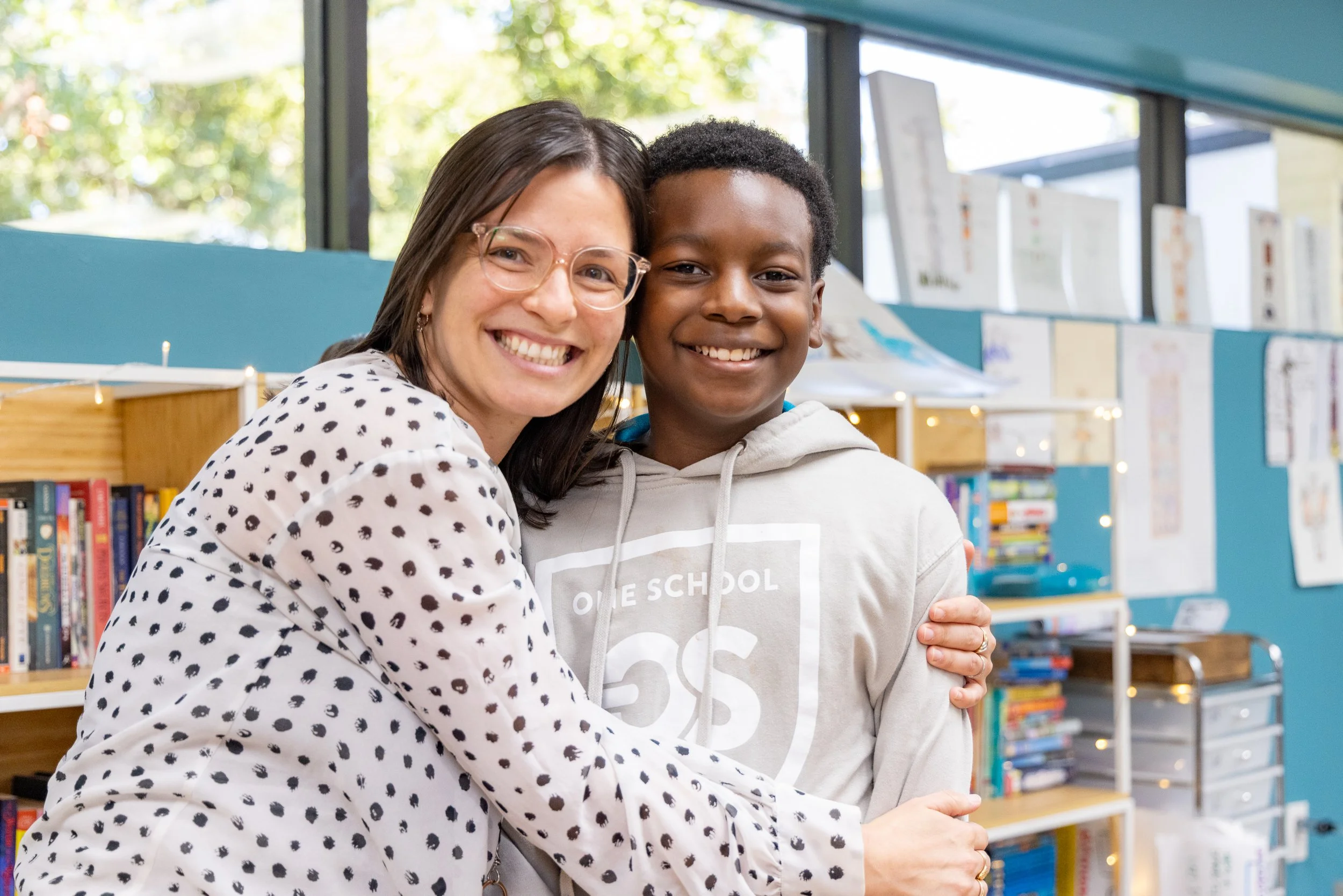 A woman and a boy smiling, hugging in a library or bookstore, with bookshelves and large windows in the background.