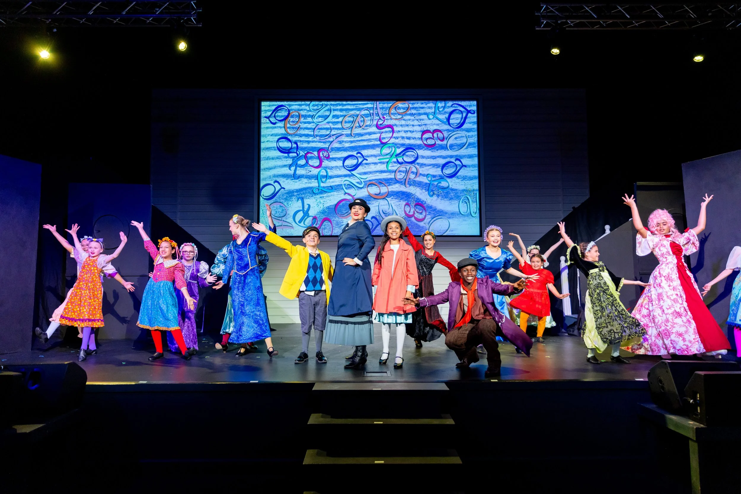 Group of children and adults dressed in colorful costumes performing a dance on a stage with a large screen displaying floating letters in the background.