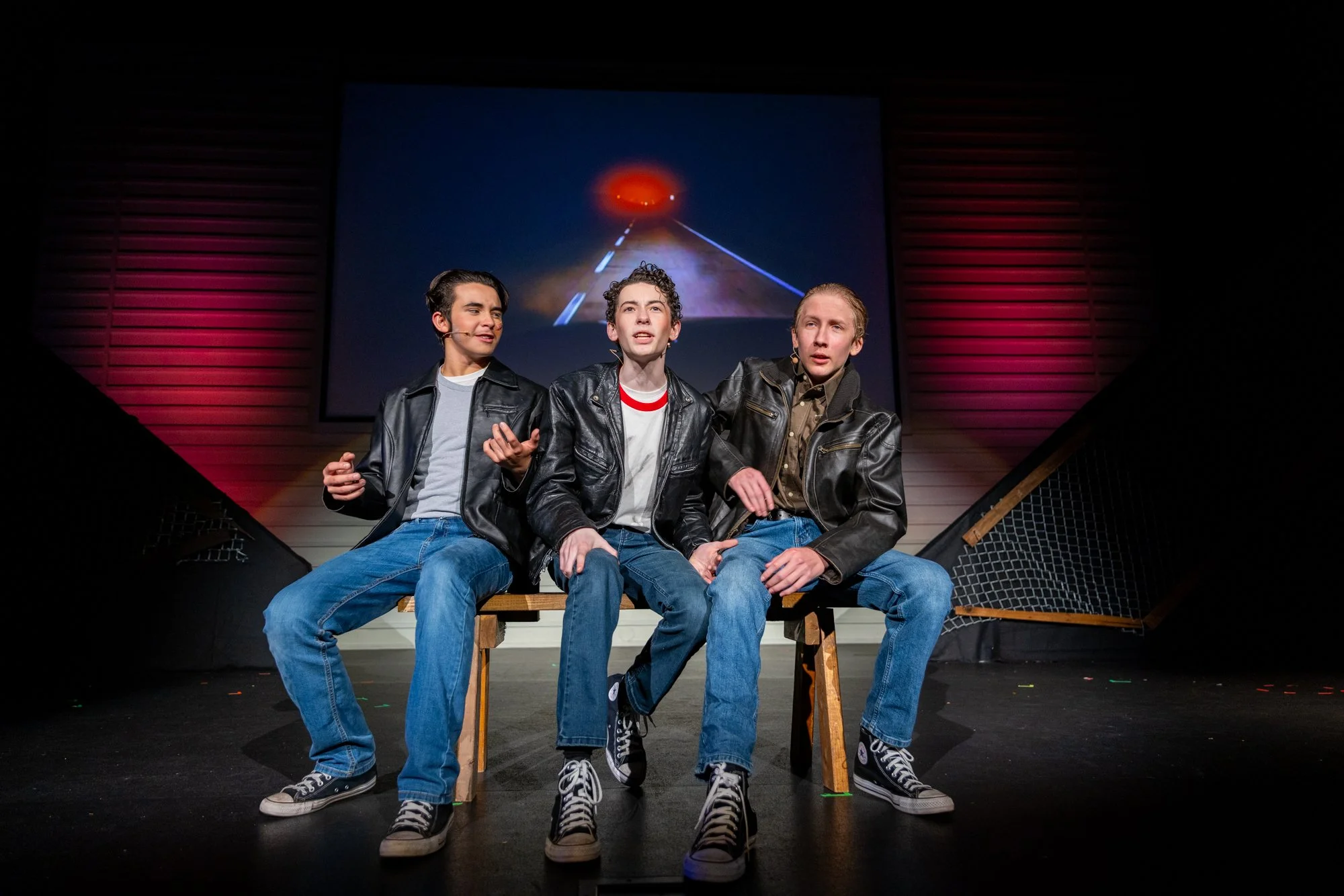 Three young boys sitting on a wooden bench on stage in a theater, appearing to perform or rehearse. They are dressed in casual clothing with leather jackets, jeans, and sneakers. The boys are speaking or singing, and there is a large screen in the background displaying a scene of a road leading into a red glowing horizon.