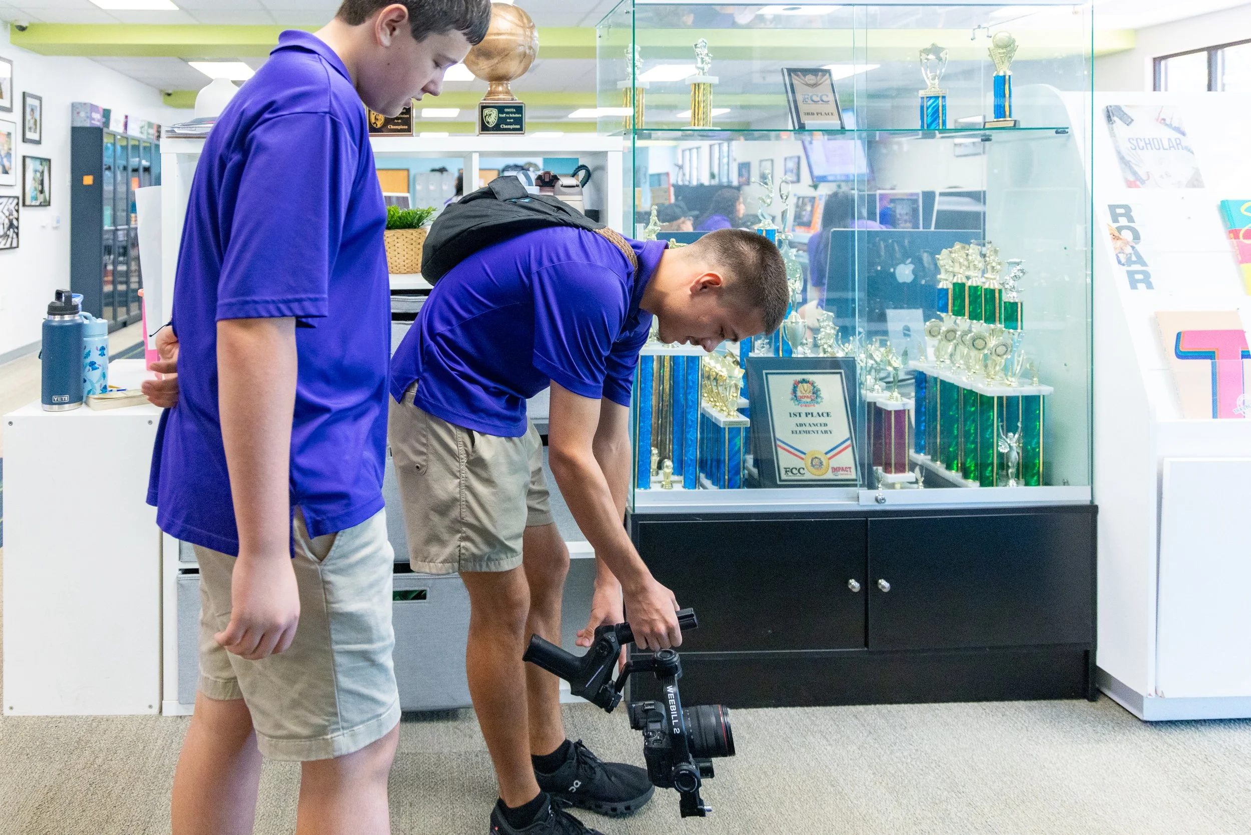 Two teenage boys in purple shirts are examining a tripod-mounted camera or binoculars in an indoor school setting, with trophy display case in the background.