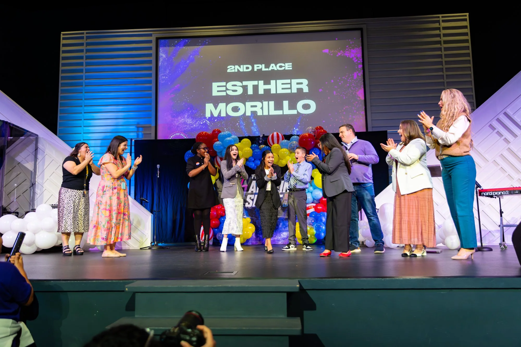 Group of people on stage celebrating second place award at an event, with balloons and a large screen displaying '2nd Place Esther Morillo'.