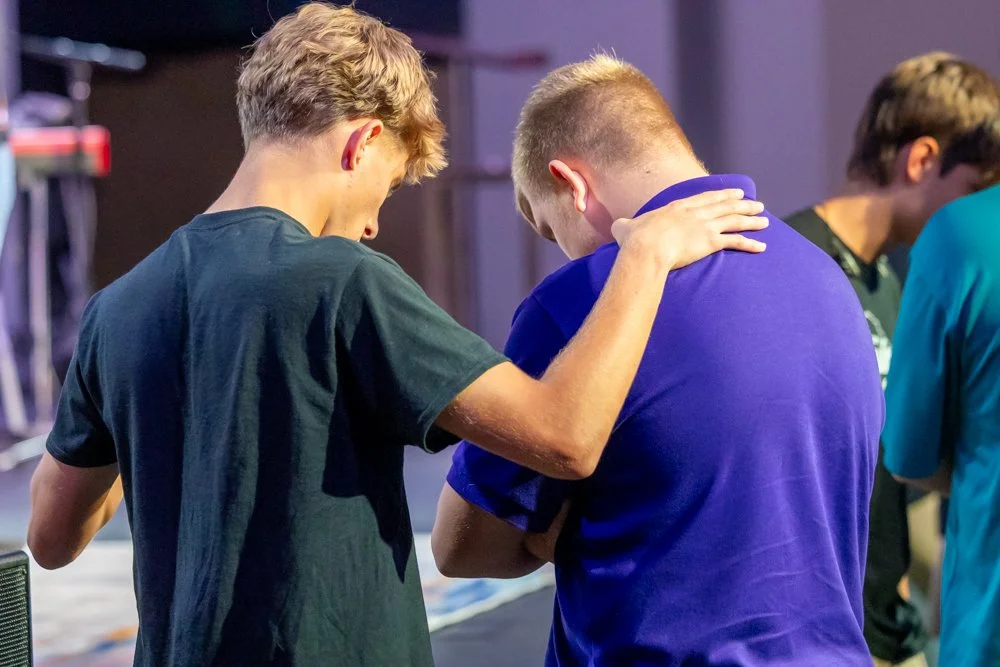 Three young men, two in black and one in blue, standing together with one man placing his arm around another's shoulder in a supportive gesture.