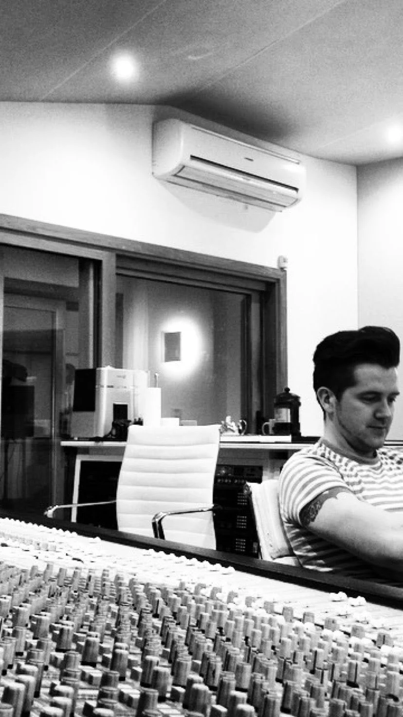 Westpoint Studios with a mixing console,  Shane Shanahan sitting in a chair, and a window showing the live room with equipment.