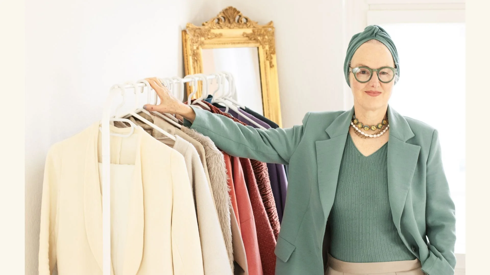 Lisa Maria Pippus, personal style expert and founder of Style Is A Language, wearing an aqua blue-green blazer and taupe trousers beside a clothing rack.