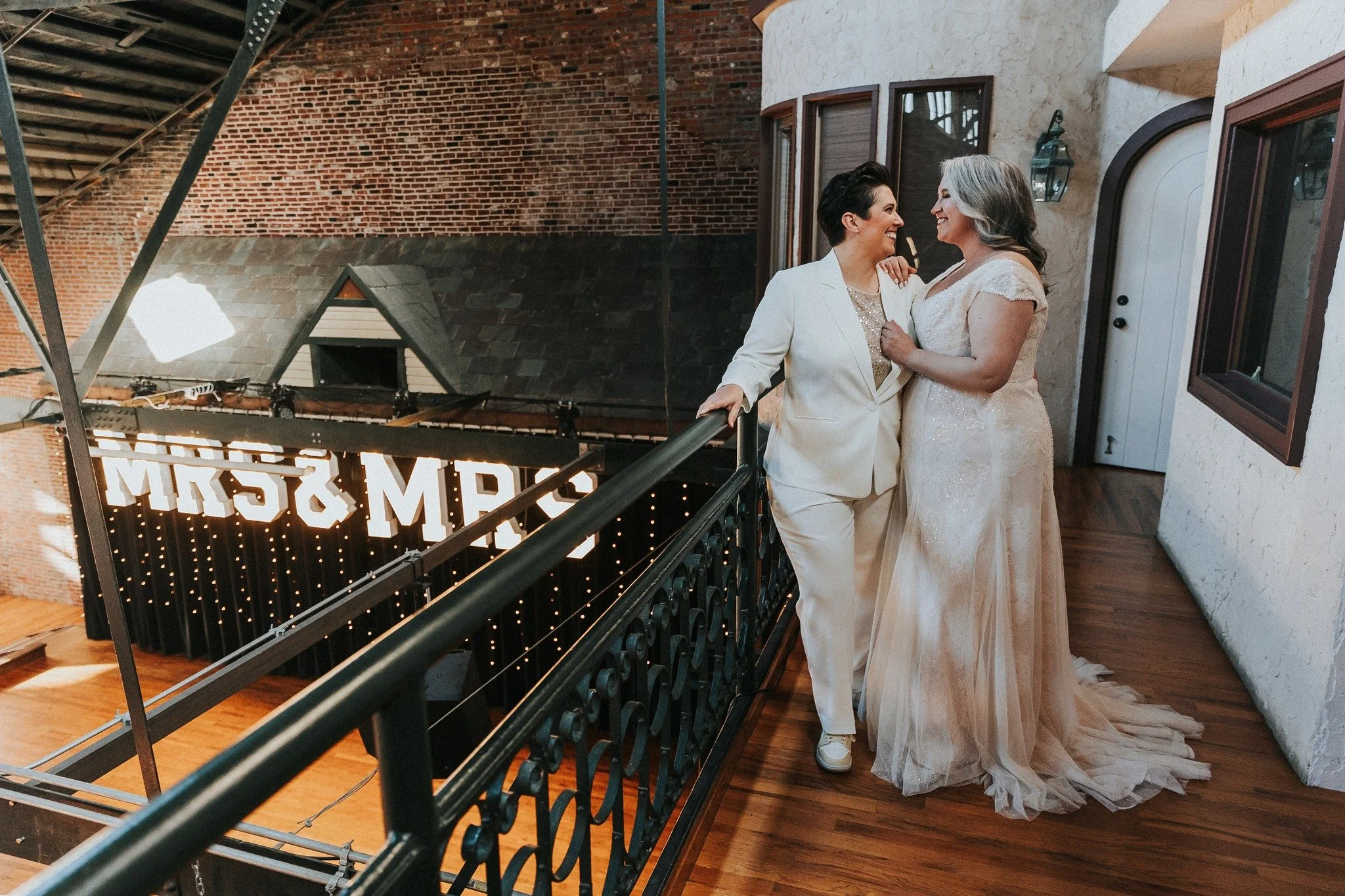 Two women, one in a white pantsuit and the other in a wedding gown, smiling and holding each other on a balcony overlooking a decorated indoor space with a neon sign that reads 'MRS & MRS'.
