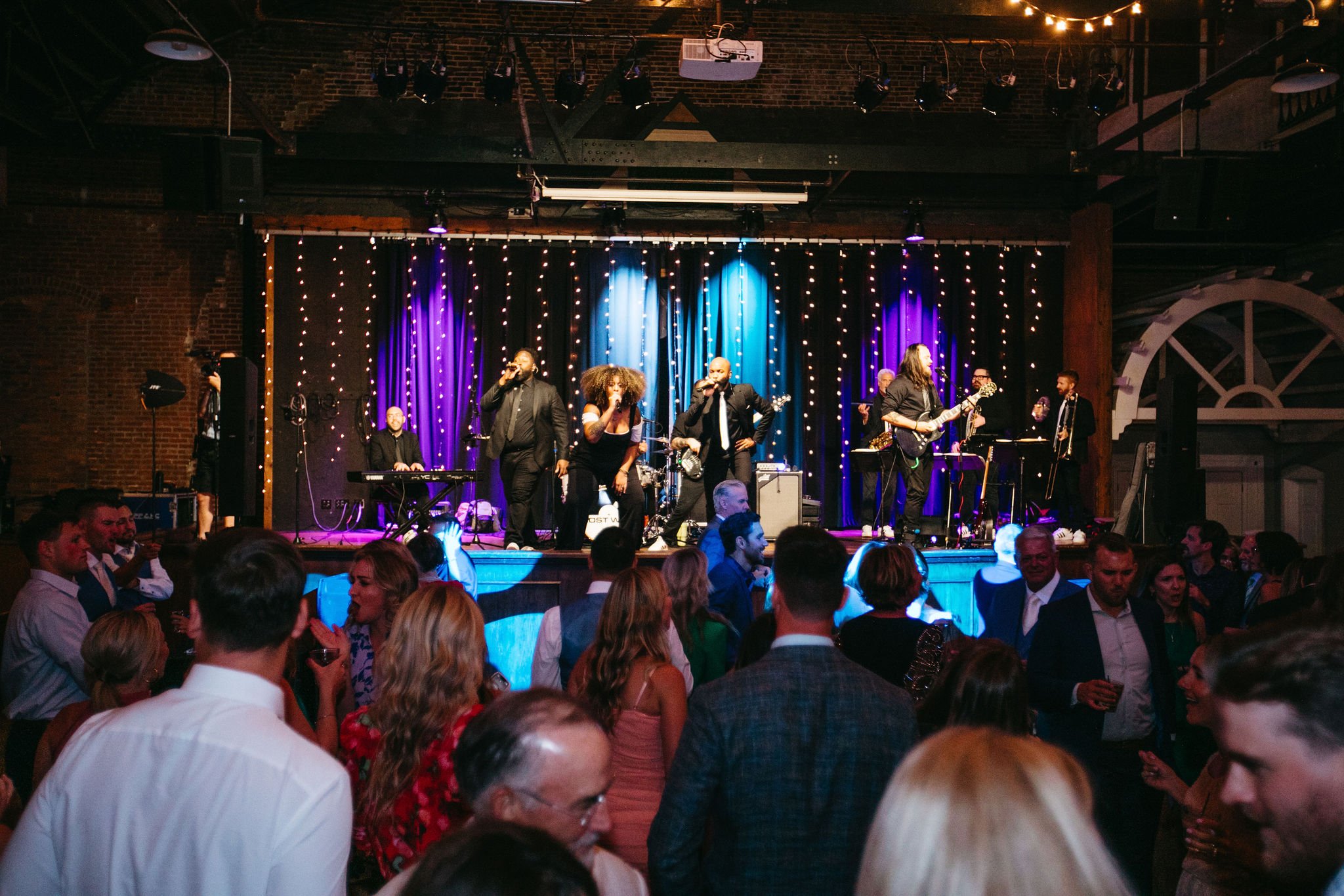 A live band performs on stage with colorful lighting, while a crowd of people dance and socialize in a lively indoor venue.