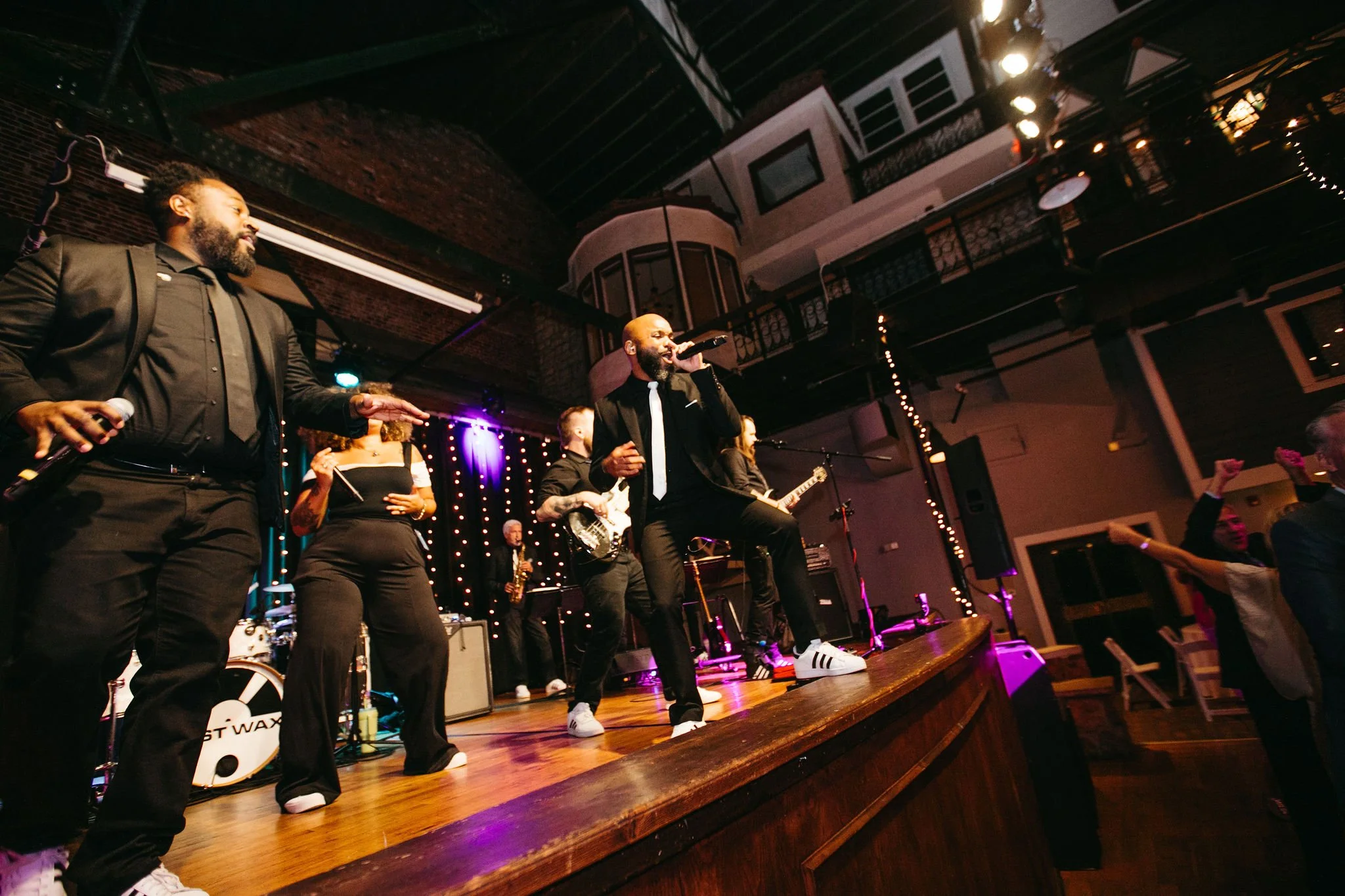 Musicians performing on stage at a concert, with one singer holding a microphone and others playing instruments, and audience members dancing.